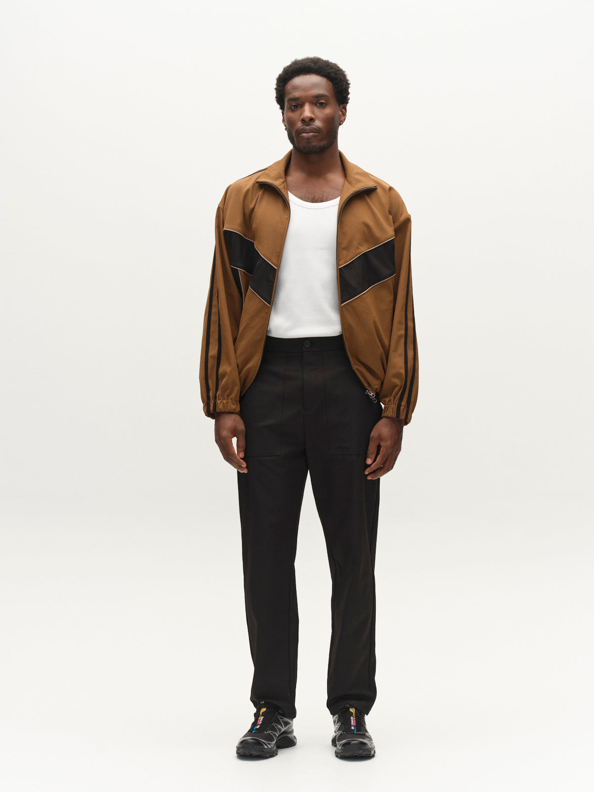 A man stands against a plain white background, wearing the Gabba GABtrack Nord Jacket in brown with black details, styled in a boxy relaxed fit. He pairs it with a white t-shirt, black sporty fabric pants, and black shoes. He faces forward with a neutral expression.