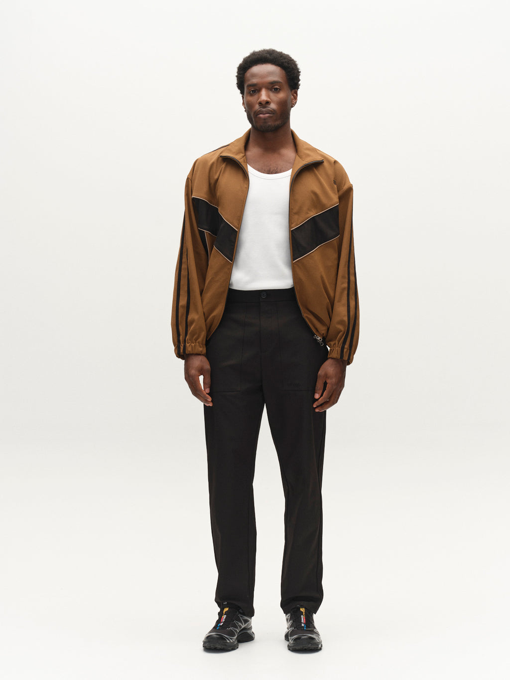 A man stands against a plain white background, wearing the Gabba GABtrack Nord Jacket in brown with black details, styled in a boxy relaxed fit. He pairs it with a white t-shirt, black sporty fabric pants, and black shoes. He faces forward with a neutral expression.