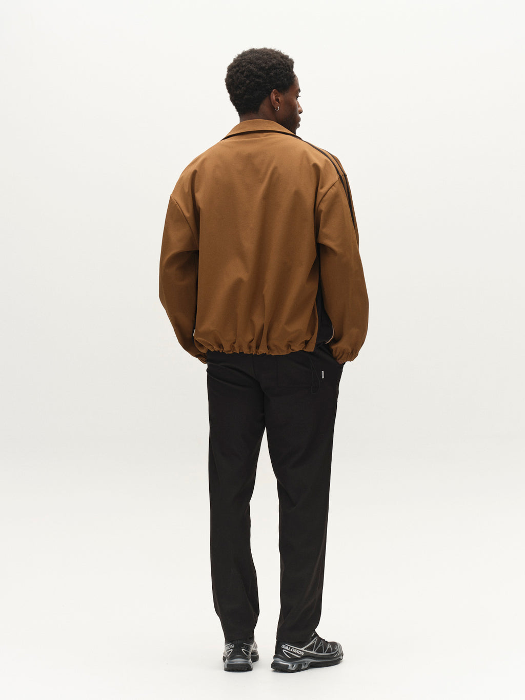 A man stands facing away, wearing the Gabba GABtrack Nord Jacket in brown with a boxy relaxed fit, paired with black pants and black sneakers, against a plain white background.