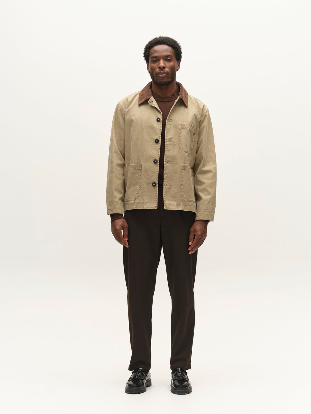 A man stands facing forward against a plain white background, wearing the Gabba GABotto Tela Outerwear - Sand Crocker, a beige washed cotton canvas jacket with a brown corduroy collar, paired with brown pants and black shoes.