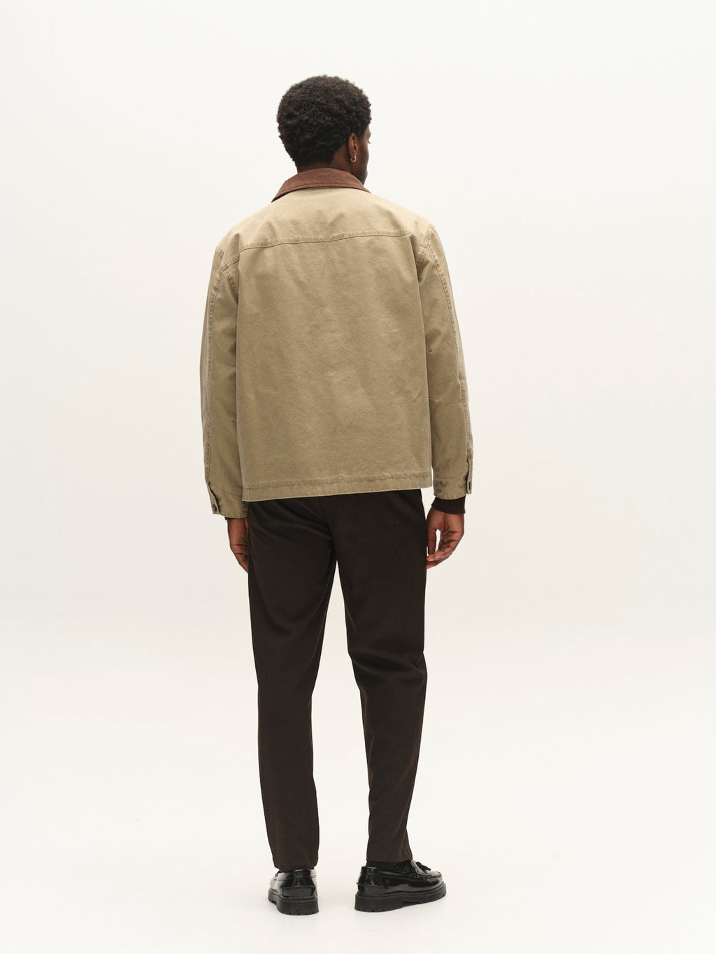 A man stands facing away, wearing the Gabba GABotto Tela Outerwear - Sand Crocker jacket with a corduroy collar, paired with dark pants and black shoes, against a plain white background.