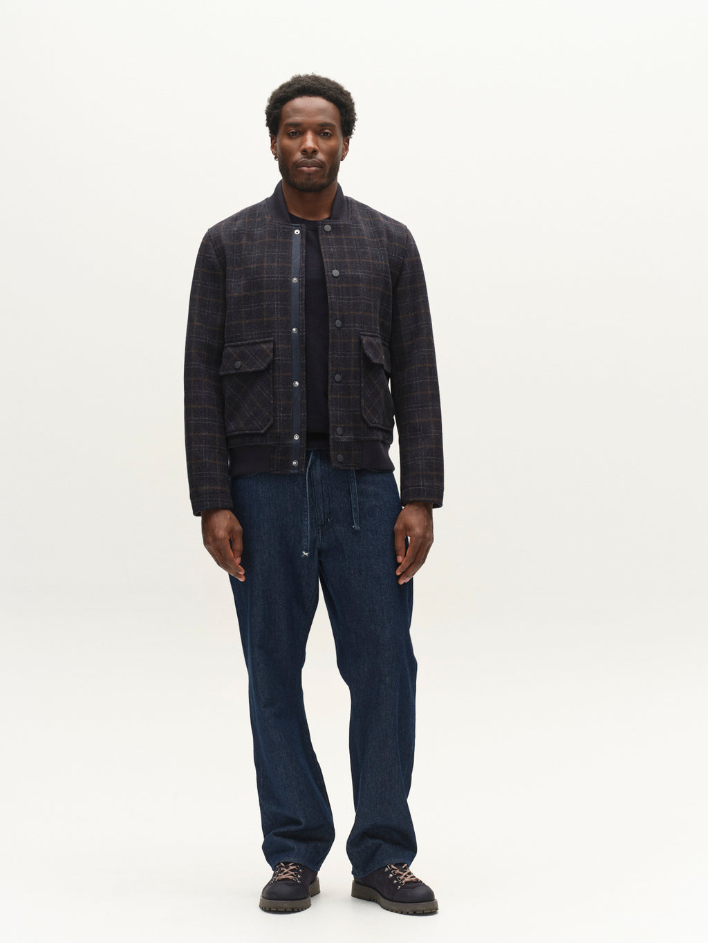 A man stands facing forward, wearing Gabba’s GABmatheo Check Bomber Jacket in Blue Check—a men’s outerwear staple—paired with a black shirt, blue drawstring pants, and black shoes, against a plain light background.