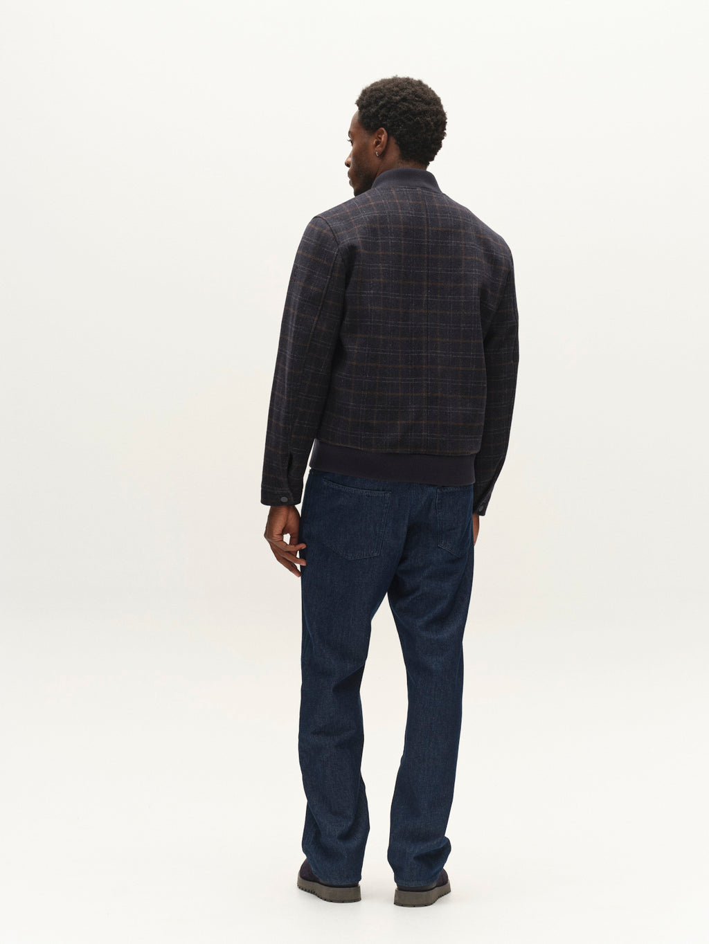 A man stands facing away, wearing the Gabba GABmatheo Check Bomber Jacket in blue check, paired with blue jeans and dark shoes against a plain white background.