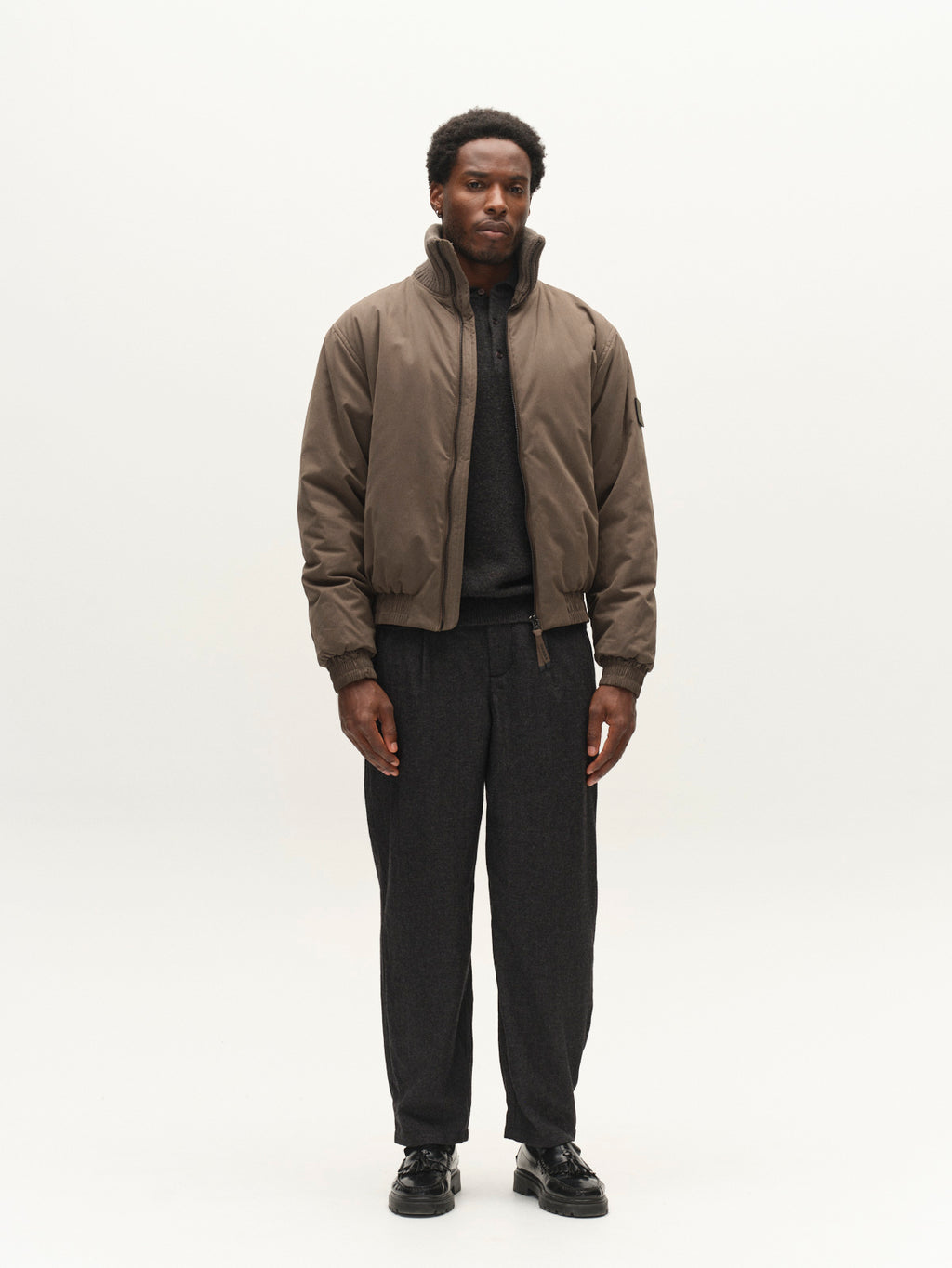 A man stands against a plain white background, wearing the Gabba GAByuri Calle Outerwear in Brown Slate—a modern bomber jacket with a boxy, relaxed fit—layered over a dark shirt and dark pants with black shoes. He faces forward with a neutral expression.
