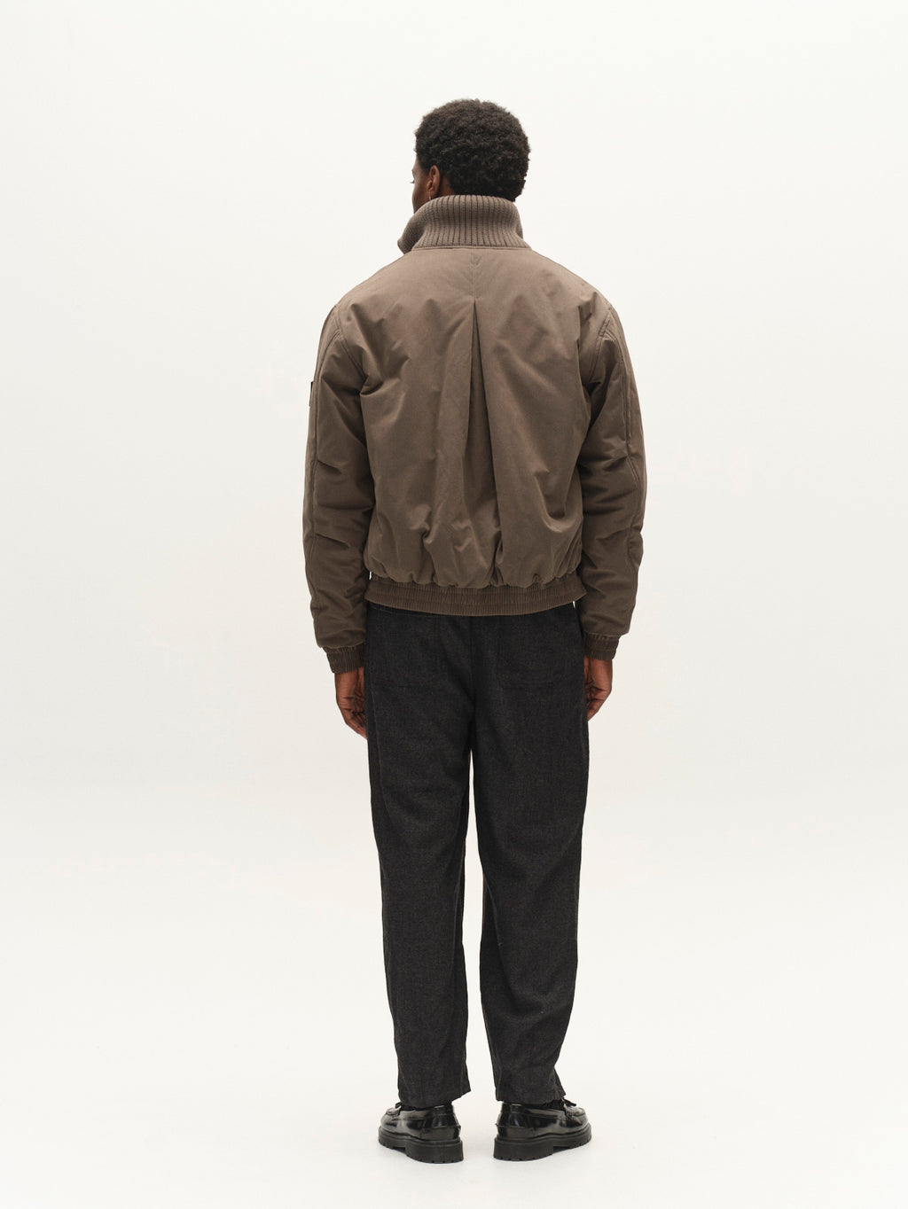 A person stands facing away from the camera, wearing the Gabba GAByuri Calle Outerwear in Brown Slate—a modern, lightly padded jacket with a boxy relaxed fit—paired with dark gray pants and black shoes against a plain, light background.
