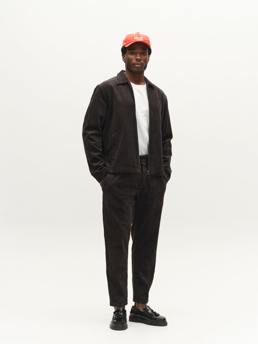 A person wearing a red cap, white t-shirt, and modern outerwear—a Gabba GABnik Zip Riglia GOTS Jacket in dark grey with matching pants—stands against a plain white background with hands in pockets. Black shoes complete the look.
