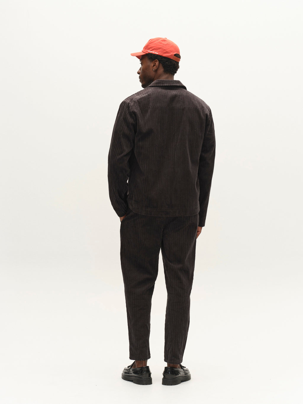 A person stands facing away against a plain white background, wearing the Gabba GABnik Zip Riglia GOTS Jacket in dark grey, paired with matching pants, black shoes, and a red cap.