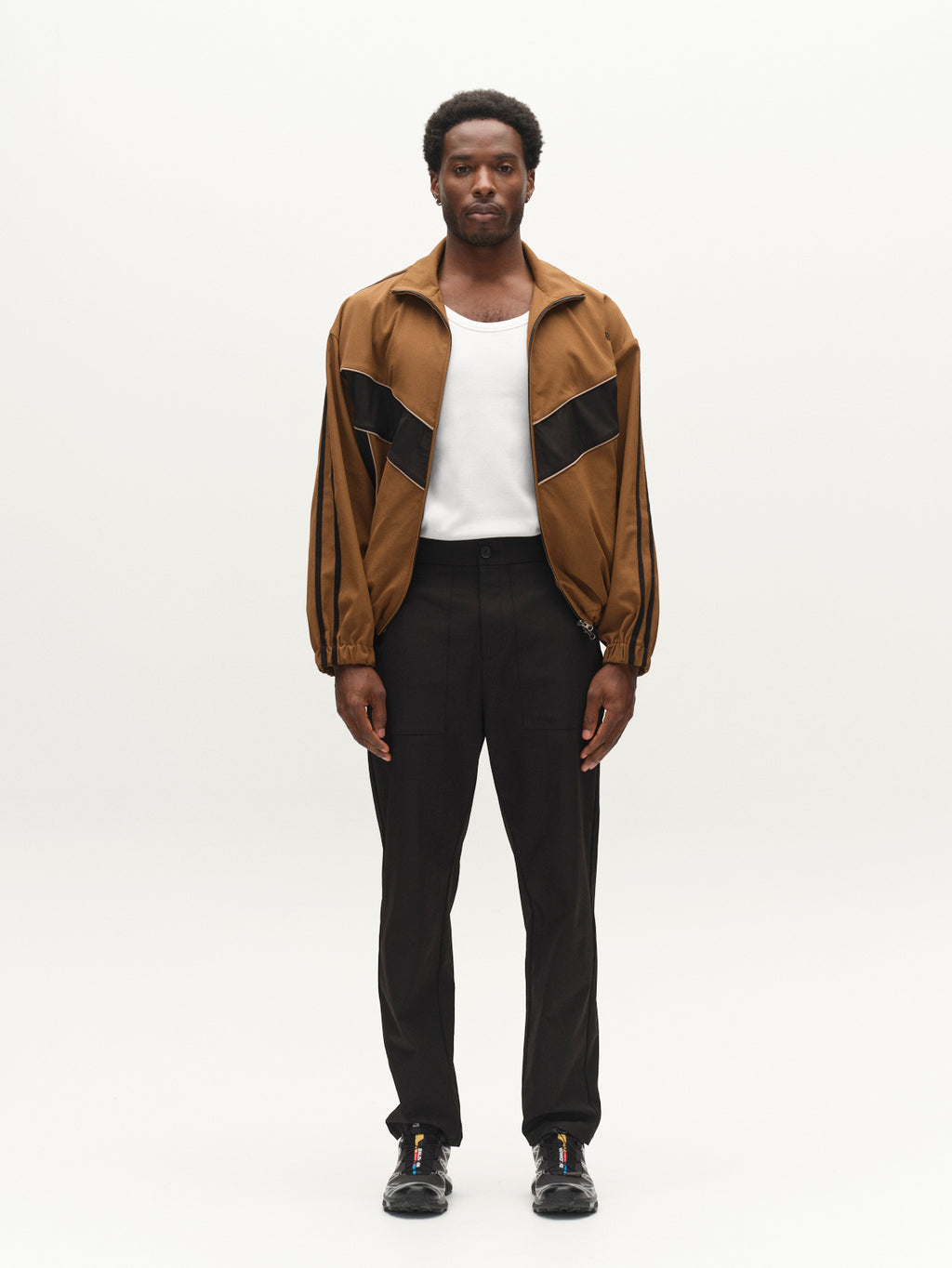 A man stands facing forward, wearing a brown zip-up jacket with black diagonal stripes crafted from lightweight technical twill, styled over a white shirt. He pairs this with the Gabba GABzeta Nord Work Pant in Meteorite and black shoes. The workwear-inspired ensemble is set against a plain white background.