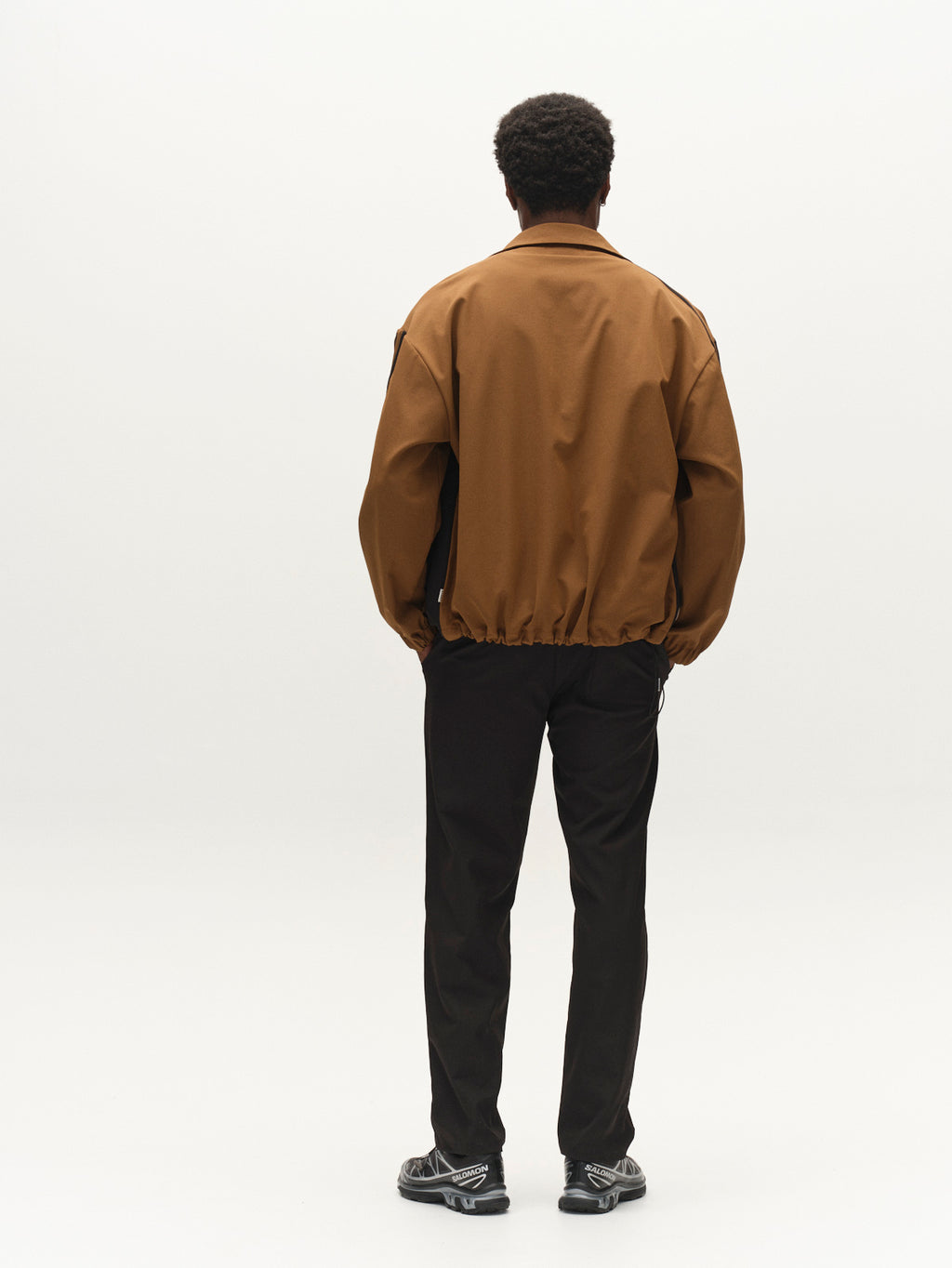 A person stands facing away, dressed in a brown jacket crafted from lightweight technical twill, paired with Gabba’s GABzeta Nord Work Pant in Meteorite and sneakers, against a plain white background.