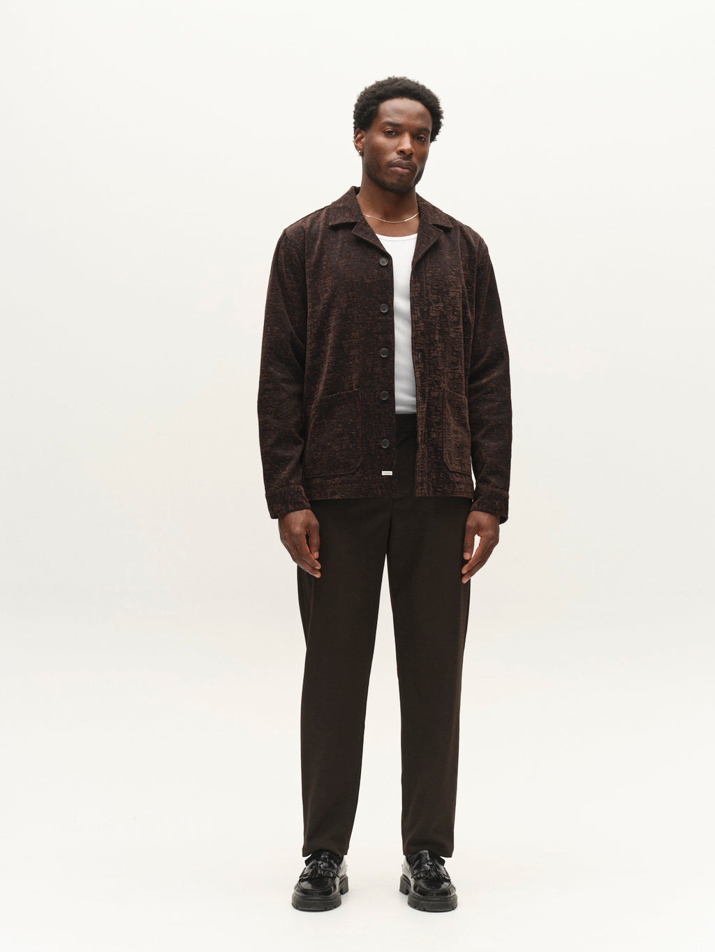 A man stands against a plain light background, wearing the Gabba GABsimone Pockt Monte LS Overshirt in Brown Slate—a dark, relaxed-fit lyocell-blend jacquard shirt layered over a white T-shirt—paired with dark trousers and black shoes. His arms rest at his sides as he looks straight ahead, embodying a contemporary statement piece from Gabba.