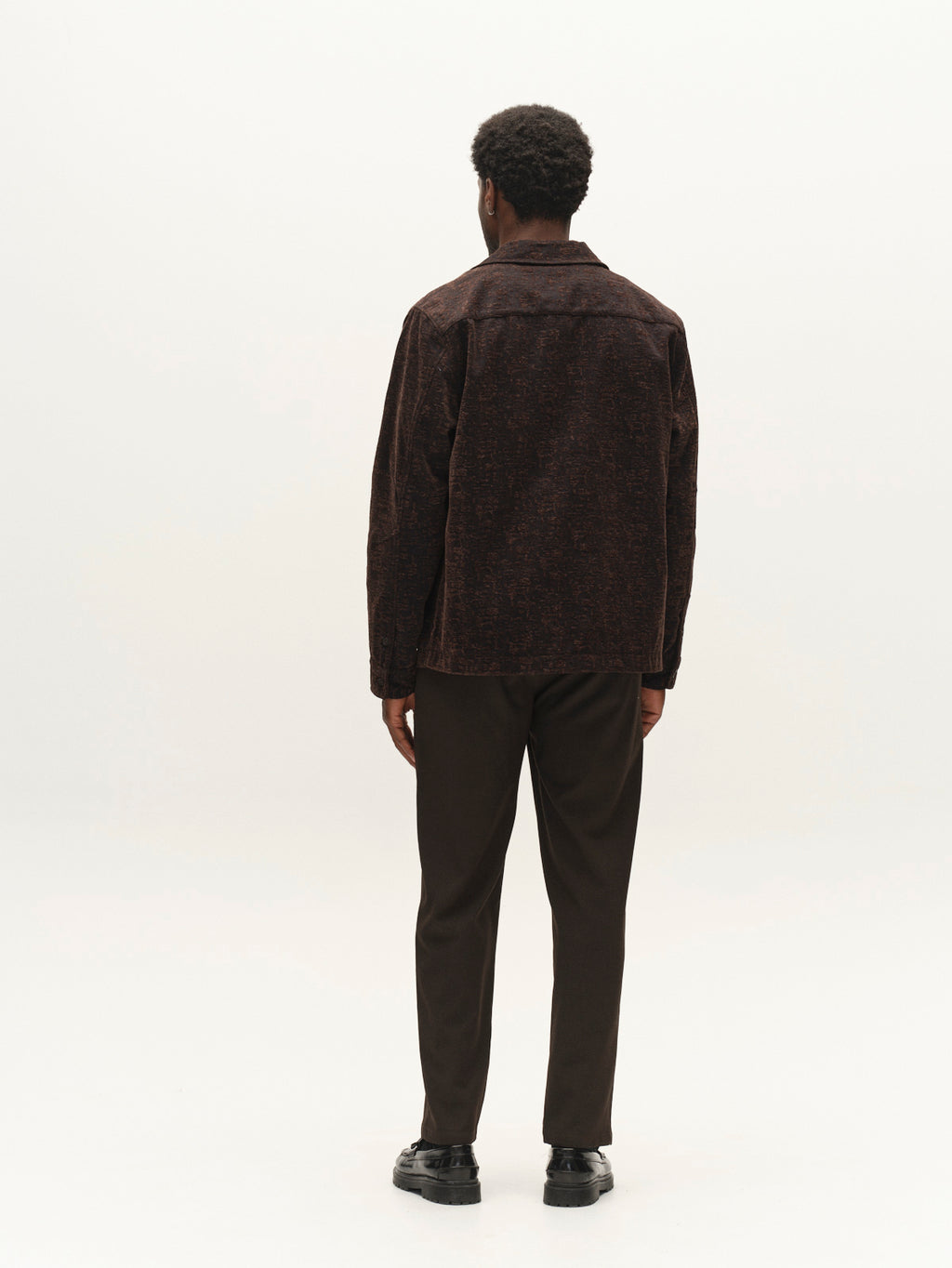 A man with short curly hair, dressed in the Gabba GABsimone Pockt Monte LS Overshirt in Brown Slate layered over a relaxed fit shirt, dark pants, and black shoes, stands facing away against a plain white background.