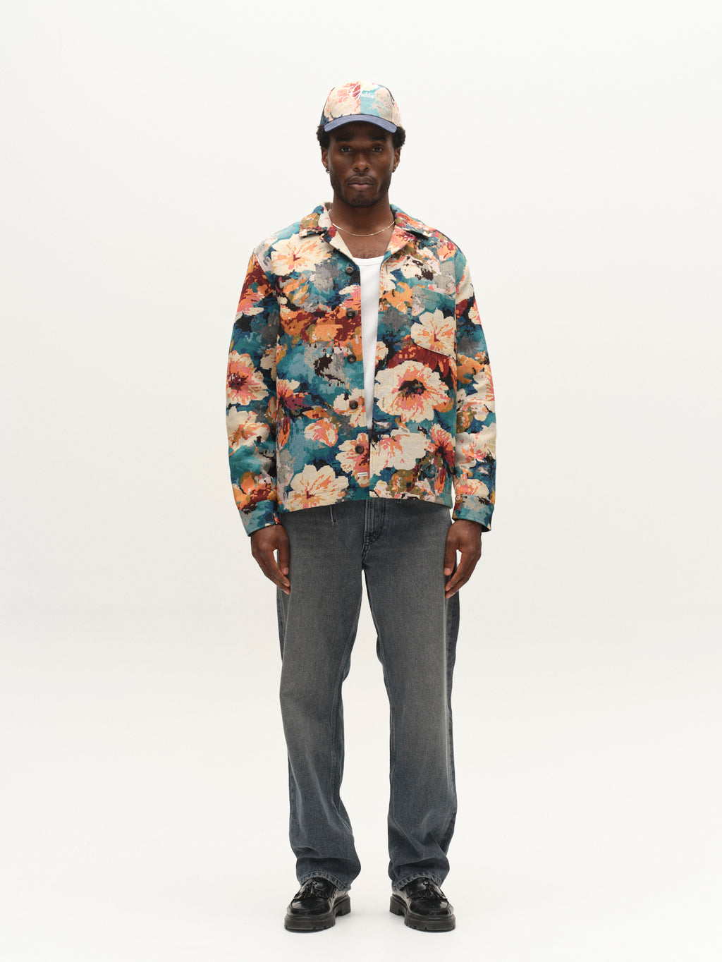 A man stands facing forward against a plain white background, wearing the Gabba GABsimone Reso Fiore LS Overshirt in a multi pattern—a relaxed fit floral piece crafted from viscose-blend jacquard fabric with a resort collar—styled with a matching cap, white shirt layered underneath, blue jeans, and black shoes.