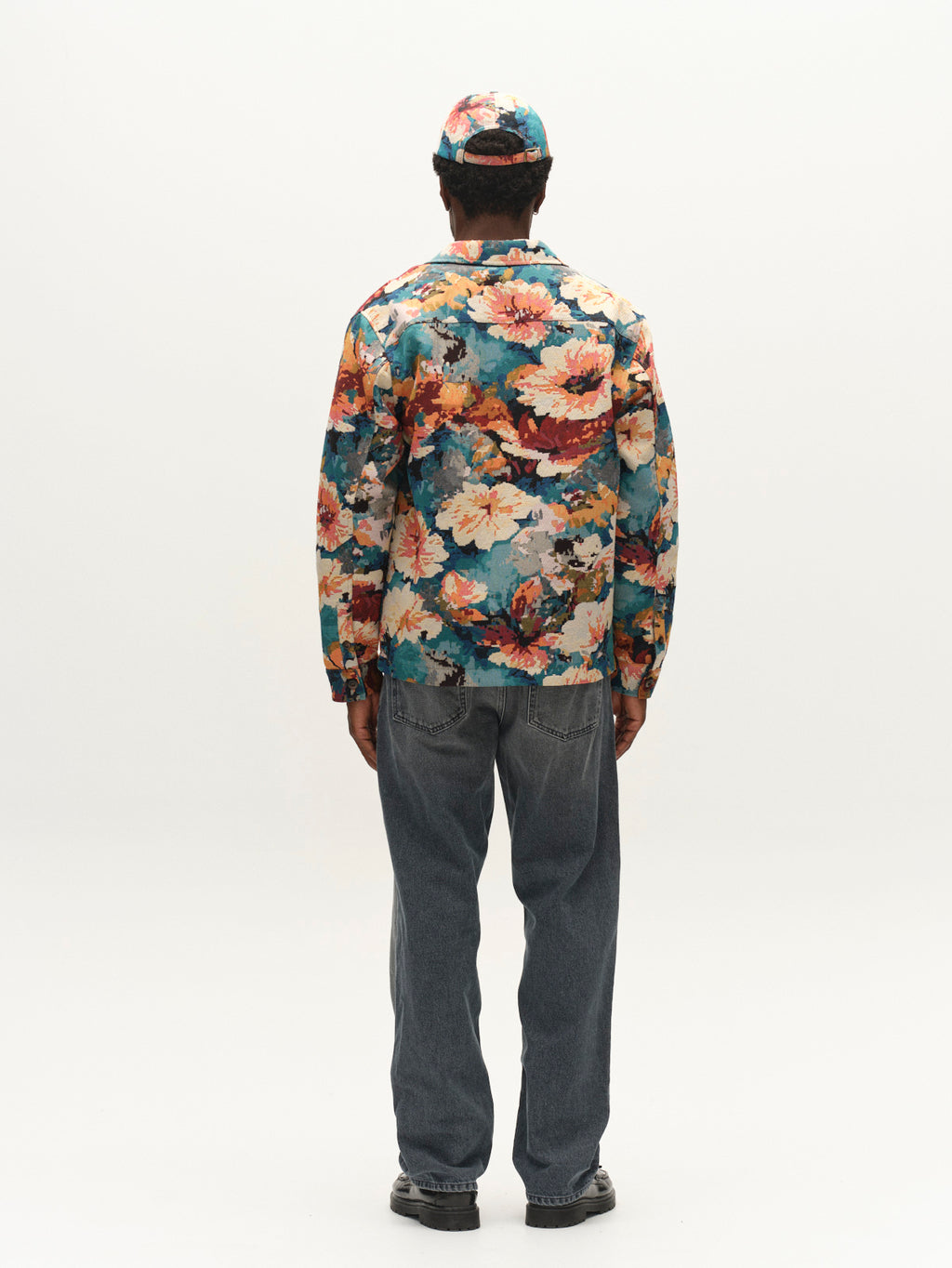 A person stands facing away, wearing the Gabba GABsimone Reso Fiore LS Overshirt in a multi-pattern floral jacquard, paired with a matching cap, blue jeans, and black shoes against a plain white background.
