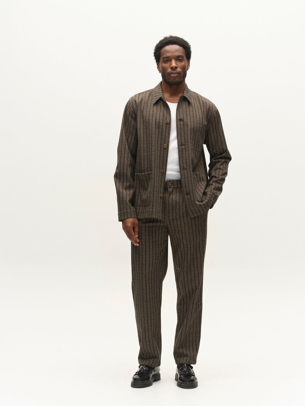 A man stands facing forward, wearing the Gabba GABlibre AX Zapies LS Overshirt in Brown Slate over a white shirt, paired with matching pinstripe trousers and black shoes, showcasing effortless lightweight layering against a plain white background.