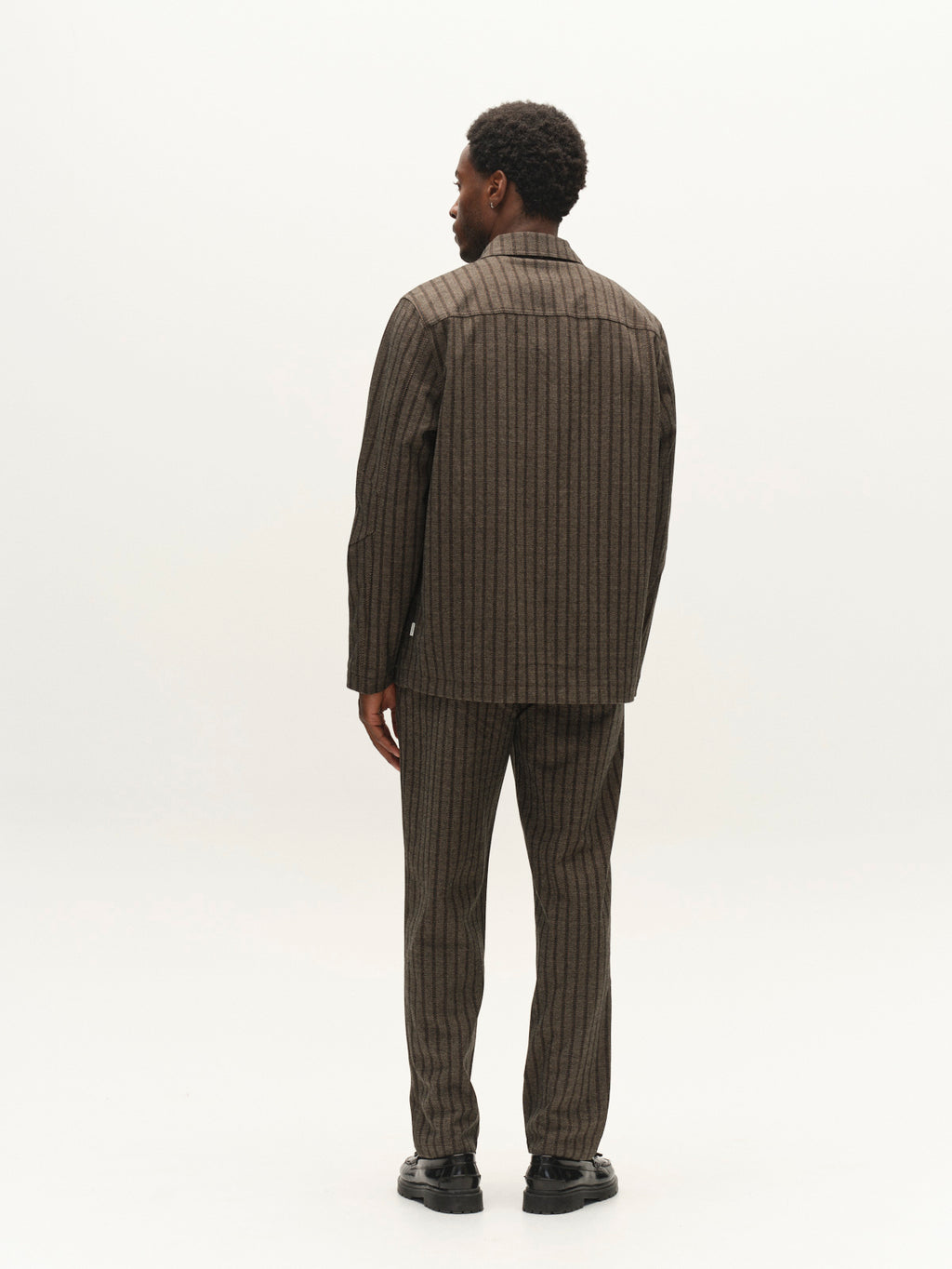 A man stands facing away from the camera, wearing a brown pinstriped regular fit suit with matching pants and black shoes, against a plain light background—perfect for lightweight layering with the Gabba GABlibre AX Zapies LS Overshirt in Brown Slate.