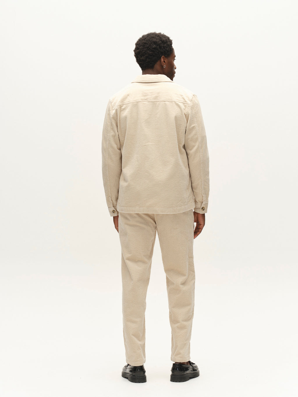 A man stands facing away, wearing the Gabba GABbeta Ibre LS Overshirt in Birch with matching pants and black shoes, against a plain white background.