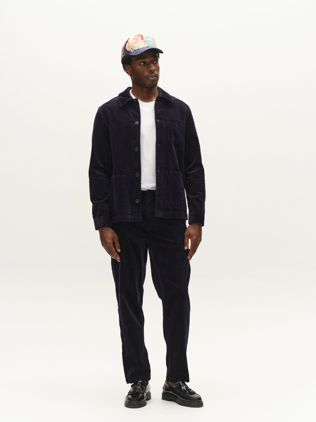 A man wearing the Gabba GABbeta Ibre LS Overshirt in Dark Navy with matching corduroy pants featuring utility pockets, a white T-shirt, black shoes, and a floral-patterned baseball cap stands against a plain white background.