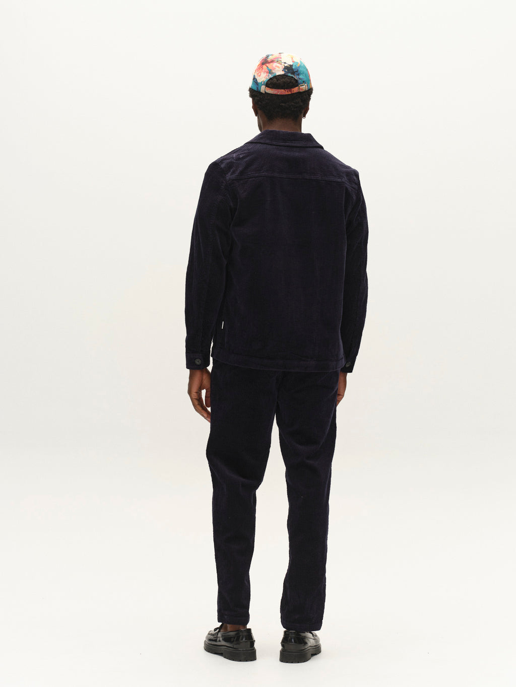 A person stands facing away from the camera, wearing the Gabba GABbeta Ibre LS Overshirt in dark navy with matching pants featuring utility pockets, black shoes, and a colorful patterned cap, set against a plain white background.