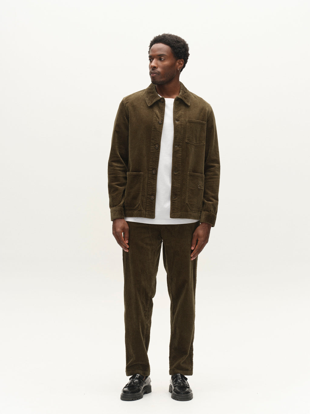 A man stands against a plain white background, wearing the Gabba GABbeta Ibre LS Overshirt in Forest Night, styled with matching pants, a white t-shirt, and black shoes. With utility pockets and an easy stretch corduroy fabric, this layering essential from Gabba combines comfort and functionality in a contemporary silhouette.