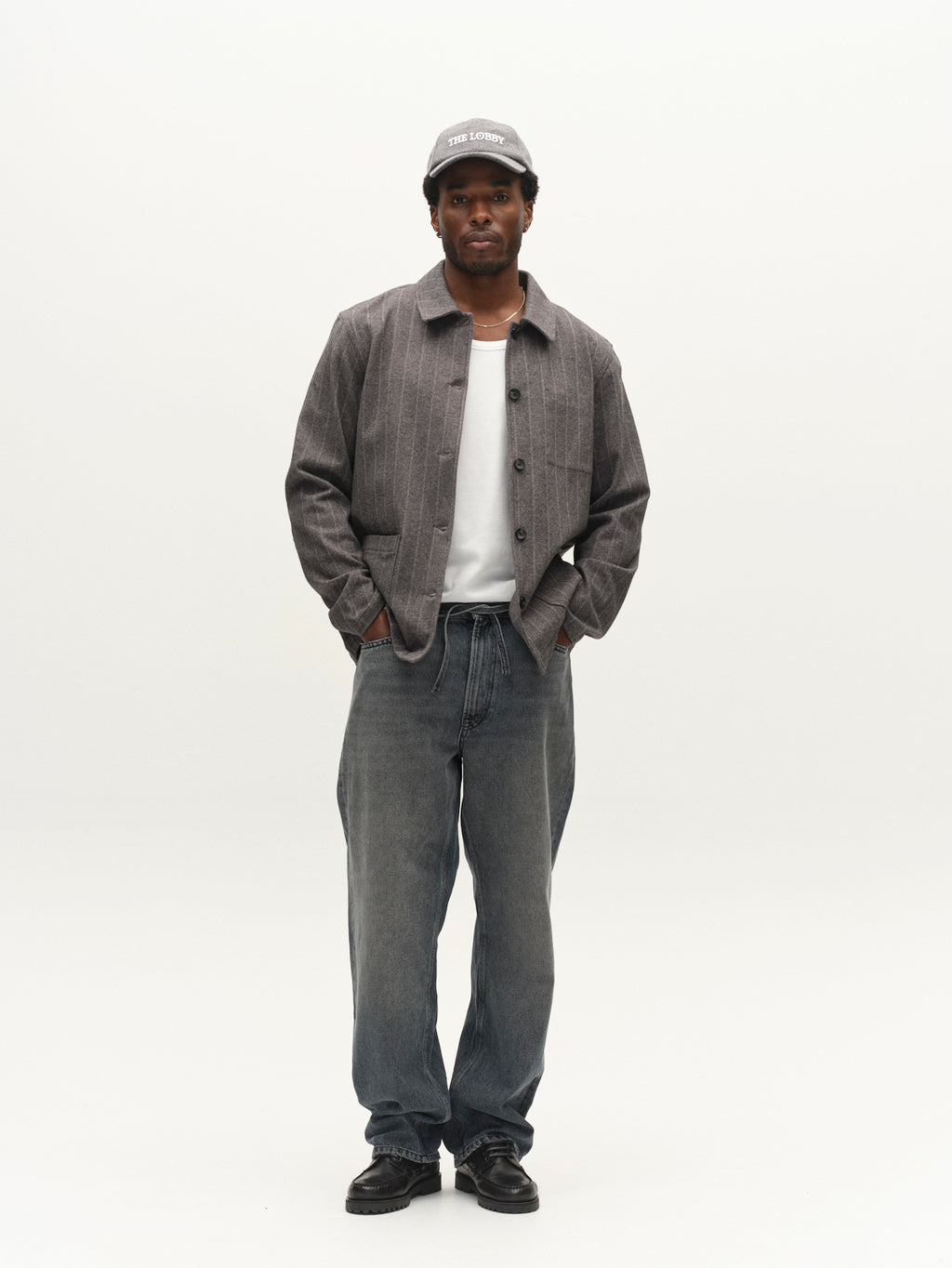 A man stands facing forward with his hands in his pockets against a plain white background, wearing the Gabba GABlibre AX Stripes LS Overshirt in dark grey, layered over a gray jacket and white shirt, paired with loose gray jeans and black shoes.