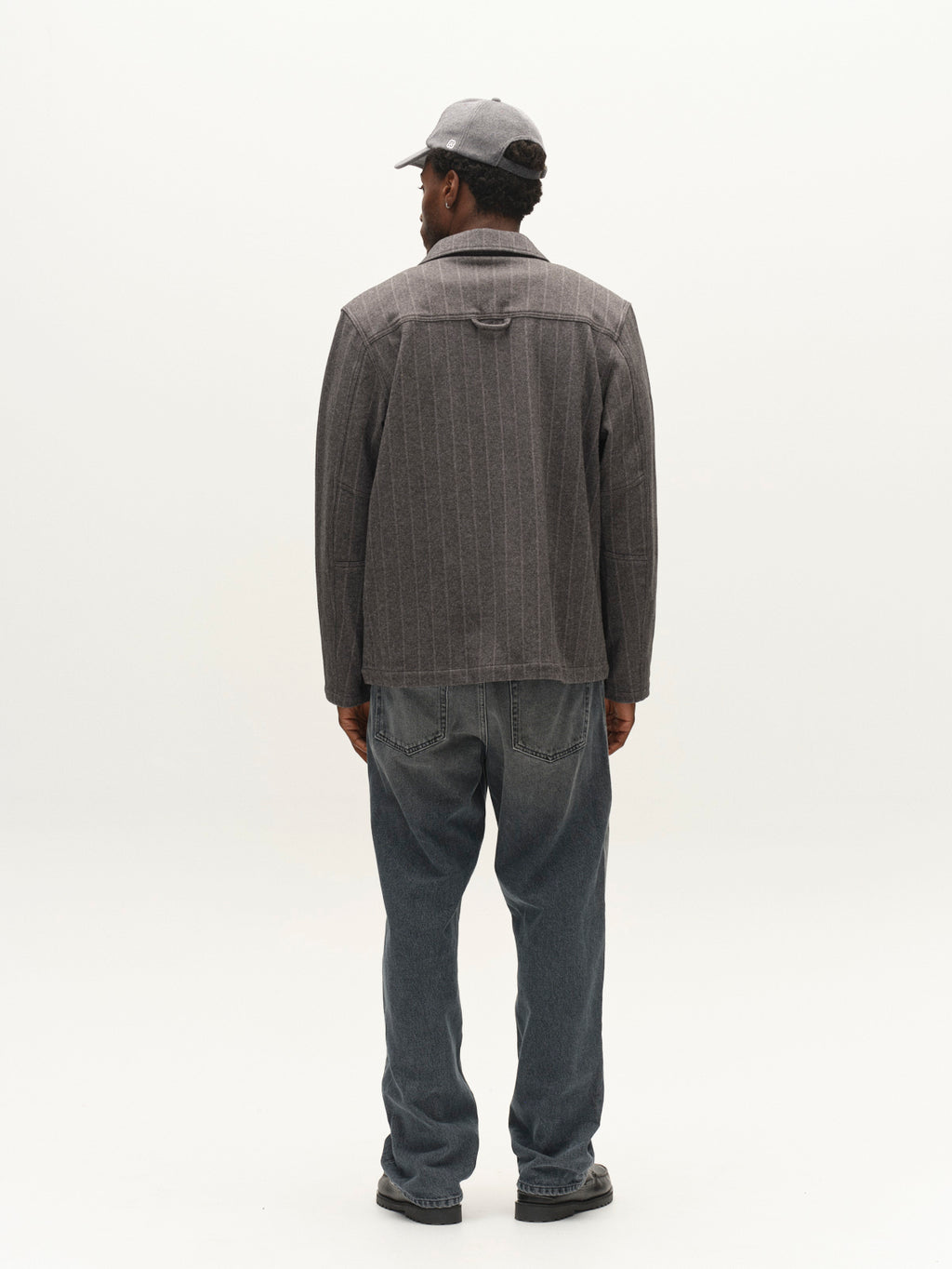 A man stands facing away, wearing the Gabba GABlibre AX Stripes LS Overshirt in Dark Grey with a boxy fit, paired with a gray cap, blue jeans, and black shoes, against a plain white background.
