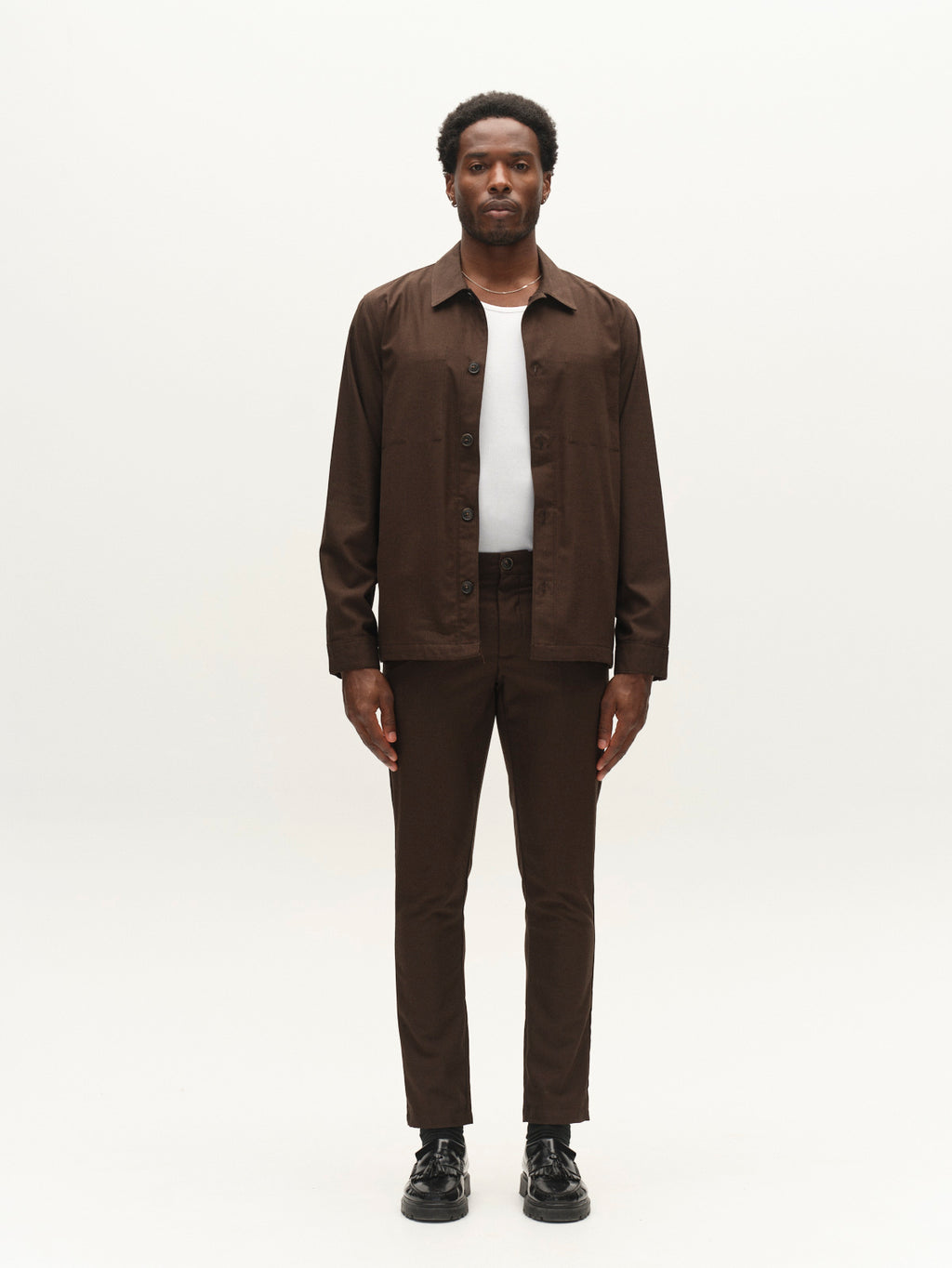 A man stands facing forward, wearing the Gabba GABtogger Base Deme LS Overshirt in Brown Slate and matching brown pants crafted from micro-pattern woven fabric, paired with a white shirt and black shoes, against a plain white background.