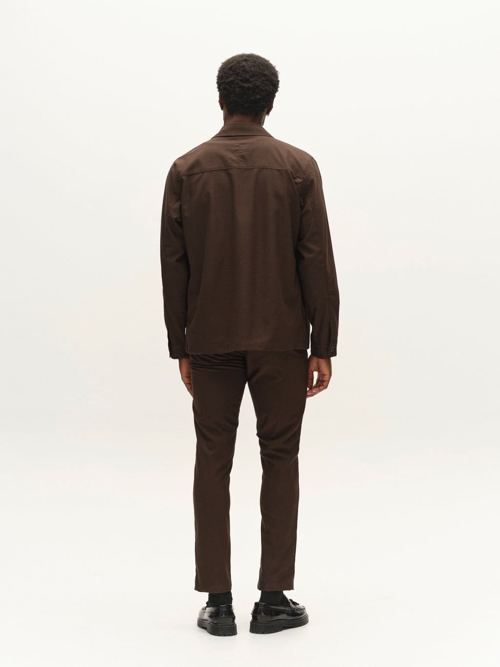 A person stands facing away from the camera, wearing the Gabba GABtogger Base Deme LS Overshirt in Brown Slate—a regular fit, long-sleeve shirt made from micro-pattern woven fabric—paired with matching brown pants and black shoes, set against a plain white background.