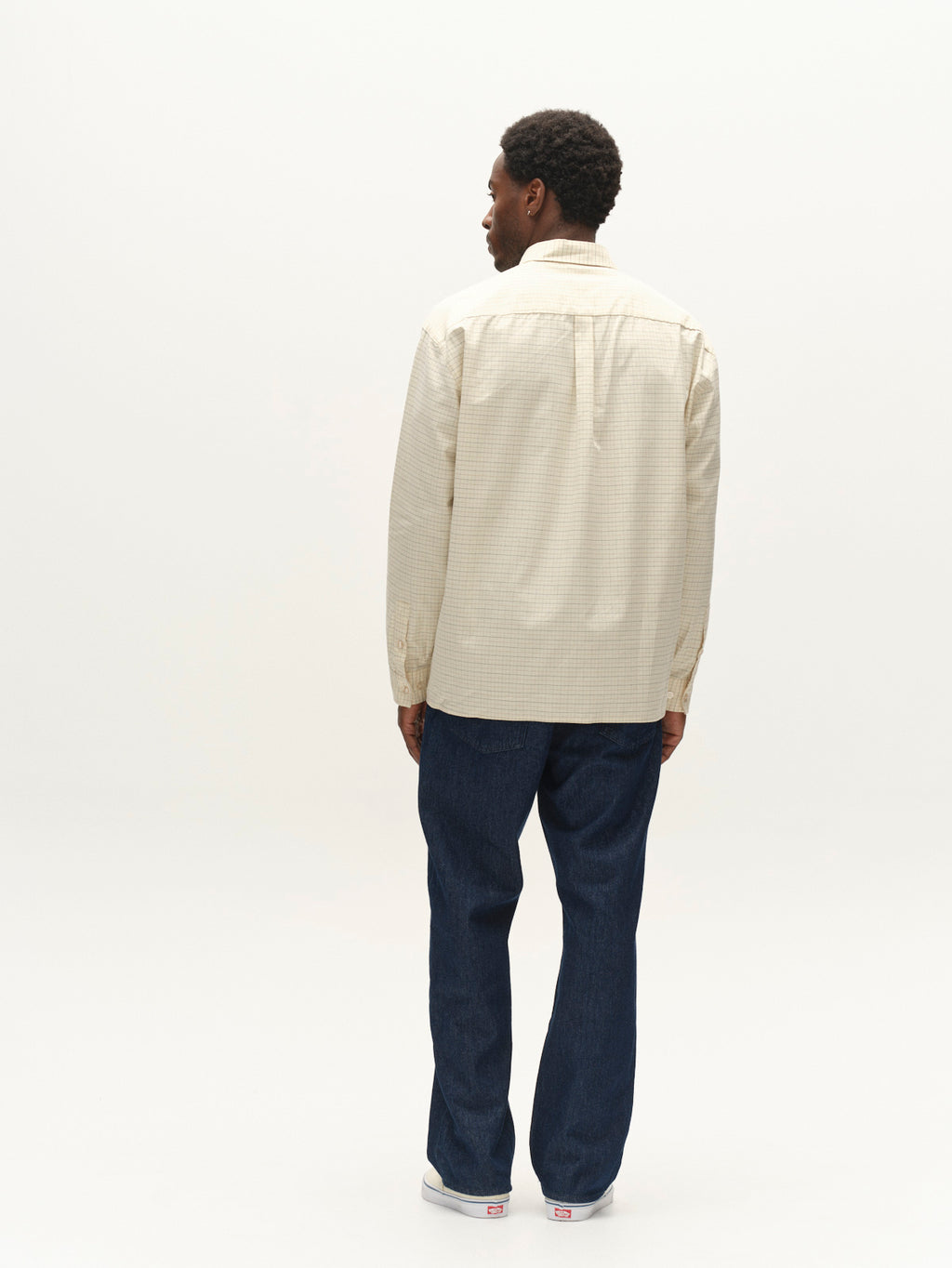 A person stands facing away from the camera, wearing the Gabba GABjens Over Indie LS Shirt in Lt. Sand—an oversized, long-sleeve shirt crafted from a cotton-blend fabric—paired with dark blue jeans and white sneakers, set against a plain white background.