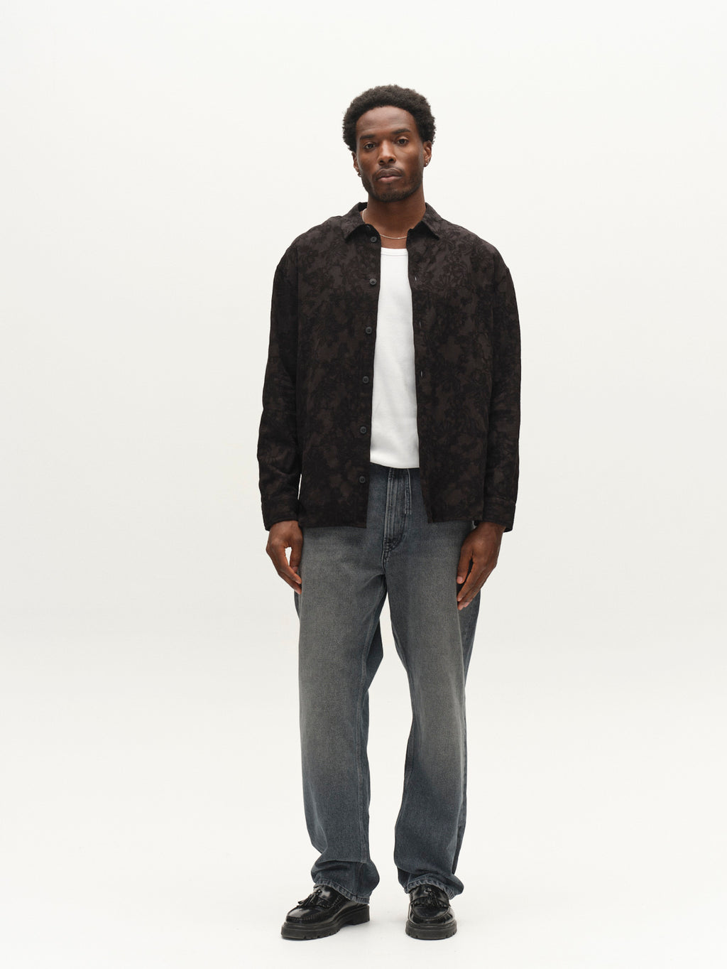 A man stands facing forward against a plain white background, wearing the Gabba GABjens Over Brocade LS Shirt in black over a white T-shirt, paired with loose blue jeans and black shoes.