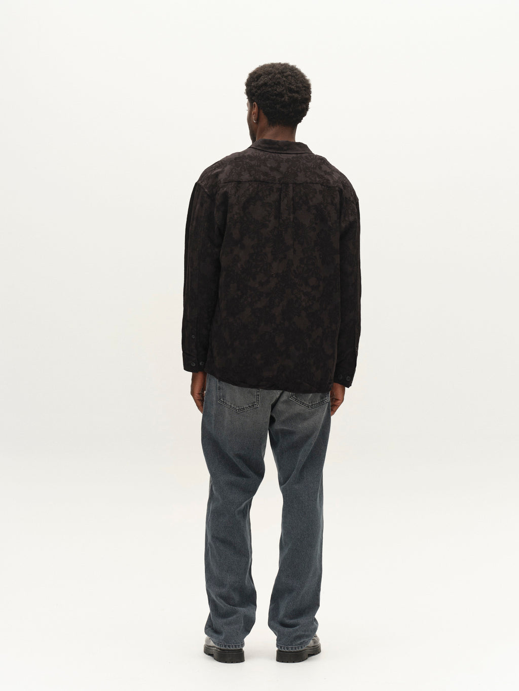 A person stands facing away, wearing the Gabba GABjens Over Brocade LS Shirt in black, styled with loose blue jeans and hands relaxed by their sides, against a plain white background.