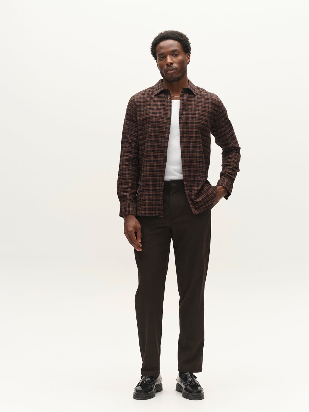 A man stands against a plain white background, wearing the Gabba GABsven Reg TX2 LS Shirt in Brown Slate over a white t-shirt, paired with dark trousers and black shoes. One hand is in his pocket as he looks calmly at the camera.