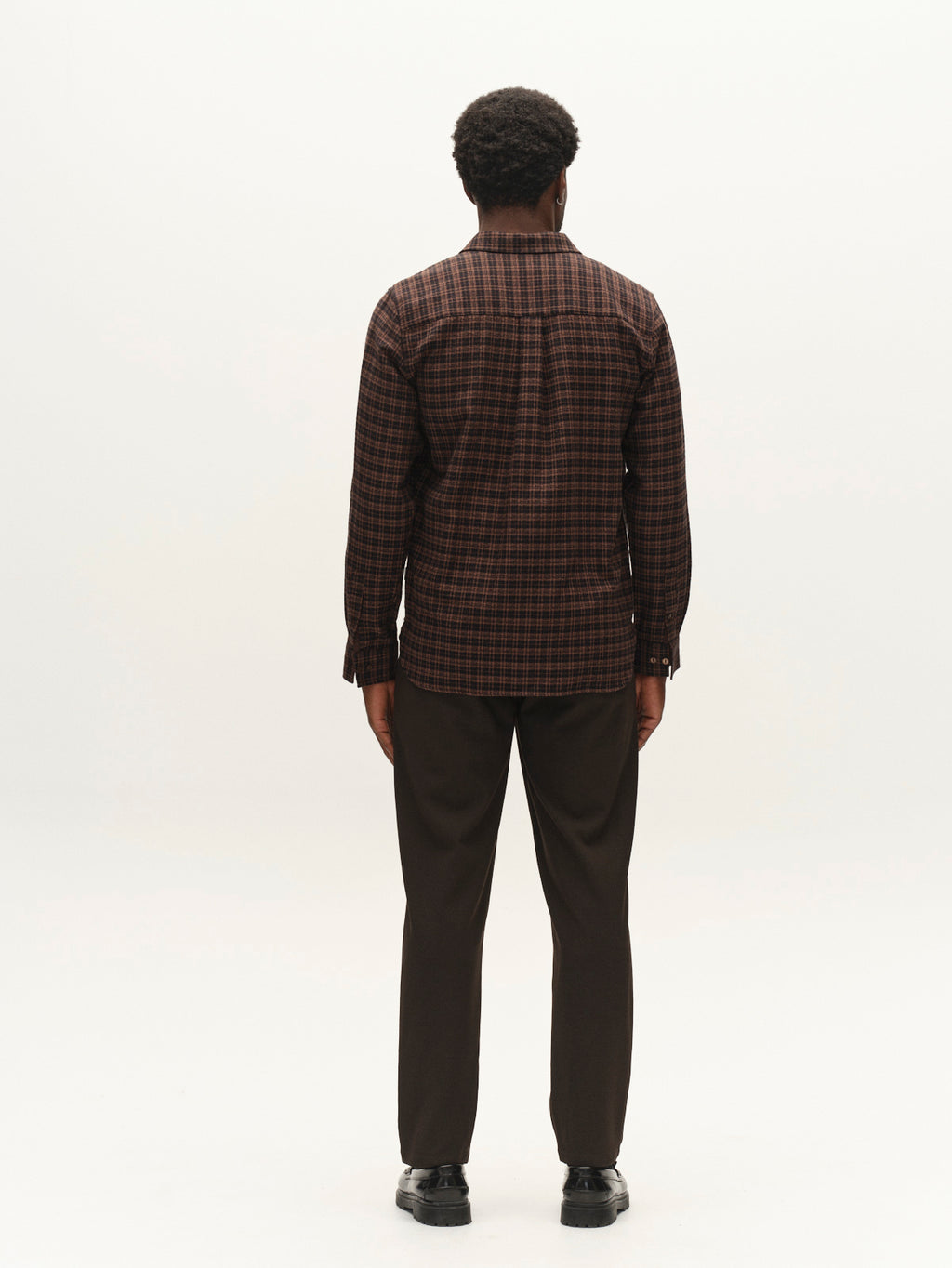 A man stands facing away from the camera, wearing the Gabba GABsven Reg TX2 LS Shirt in Brown Slate—a yarn-dyed cotton regular-fit shirt with a checked texture—paired with dark pants and black shoes, set against a plain white background.