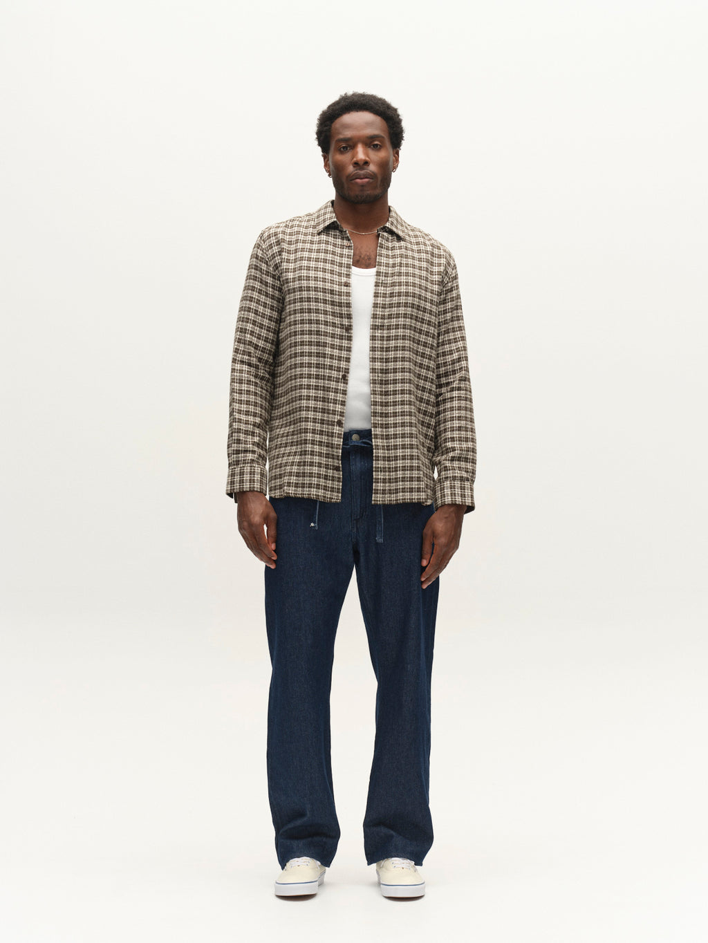 A man stands against a plain white background, wearing the Gabba GABsven Reg TX2 LS Shirt in Forest Night—a regular fit, yarn-dyed cotton shirt with a checked texture—styled over a white T-shirt, blue jeans, and white sneakers. The subtle detailing of the shirt adds interest to his outfit. He faces forward with a neutral expression and his arms by his sides.