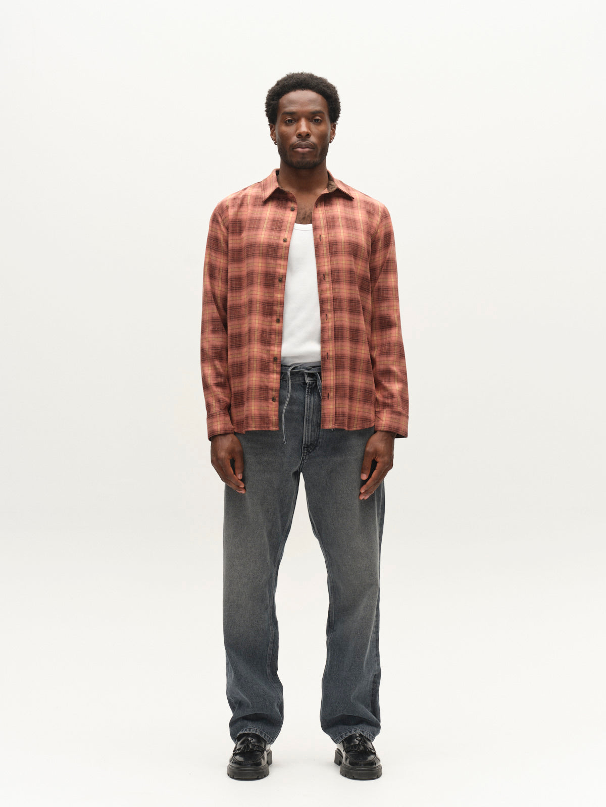 A man stands facing forward against a plain white background, wearing the Gabba GABsven Reg F1097 LS Shirt in Brick Red over a white top, paired with loose gray drawstring jeans made from a stretch-viscose blend, and black shoes.