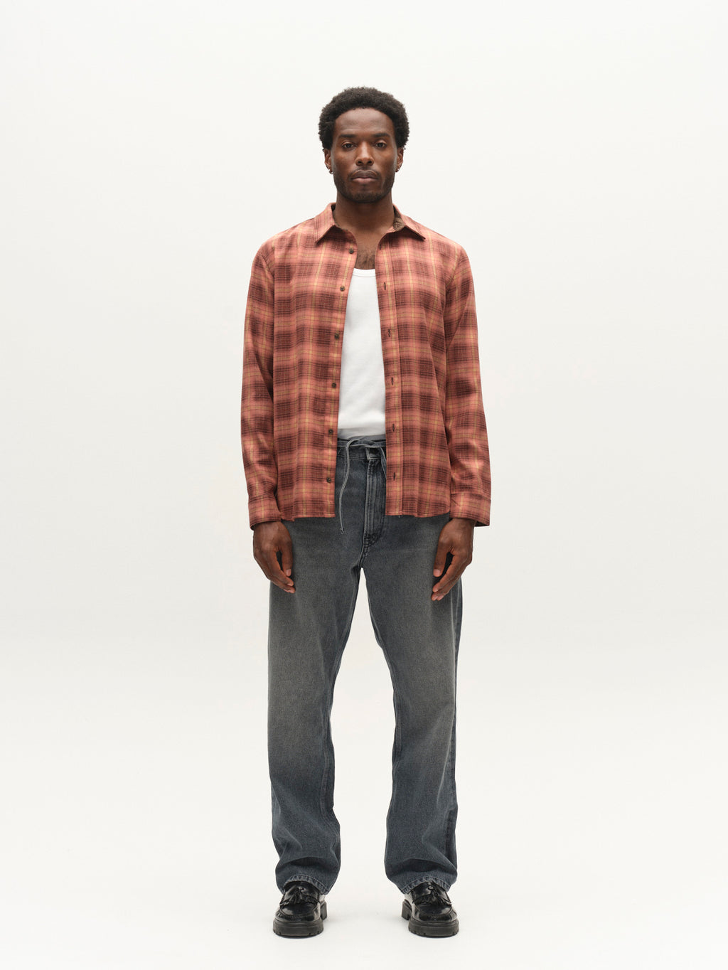 A man stands facing forward against a plain white background, wearing the Gabba GABsven Reg F1097 LS Shirt in Brick Red over a white top, paired with loose gray drawstring jeans made from a stretch-viscose blend, and black shoes.
