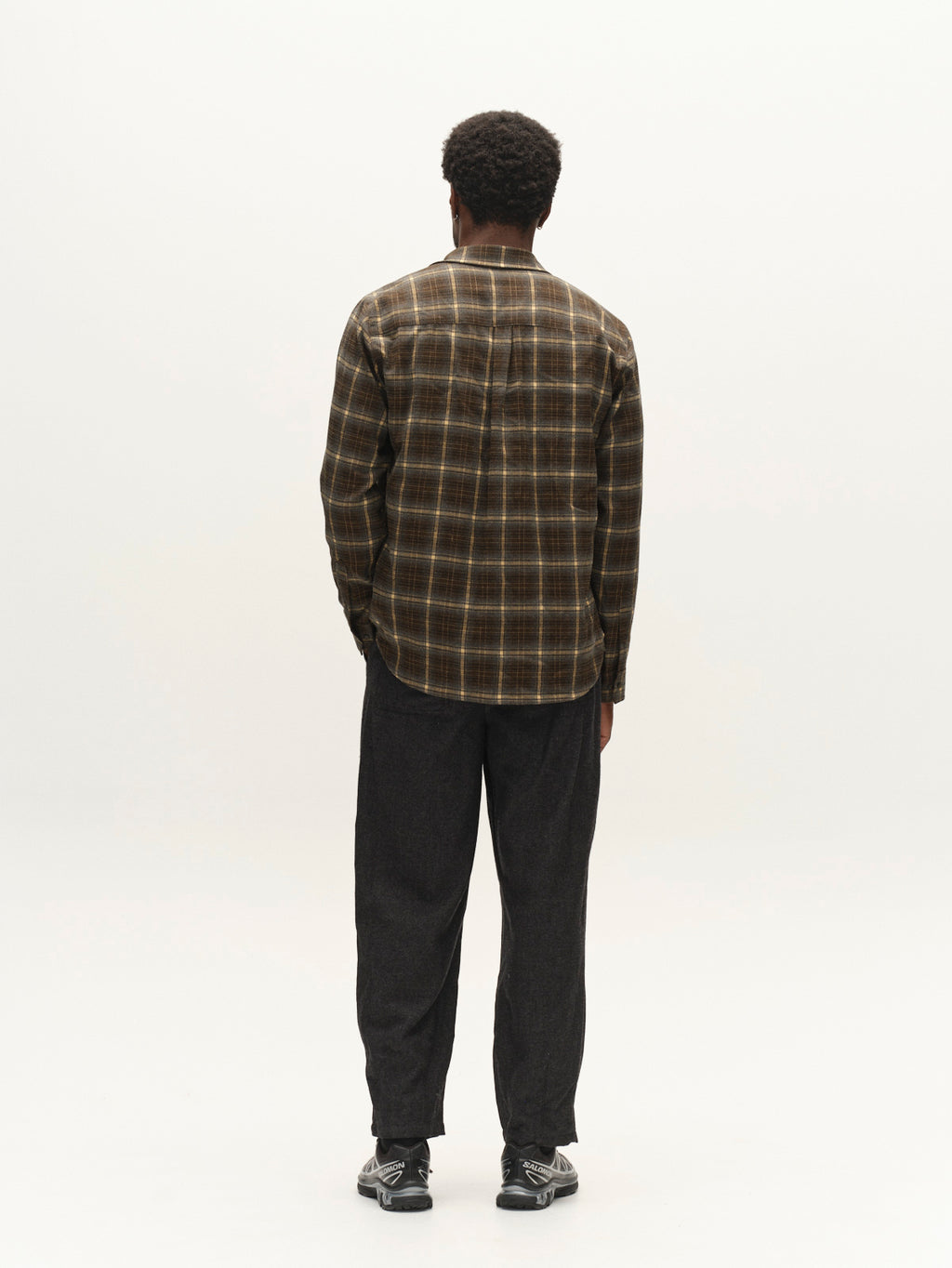 A person stands facing away, wearing the Gabba GABsven Reg F1097 LS Shirt in Forest Night, paired with dark pants and black sneakers. The background is plain and light-colored.