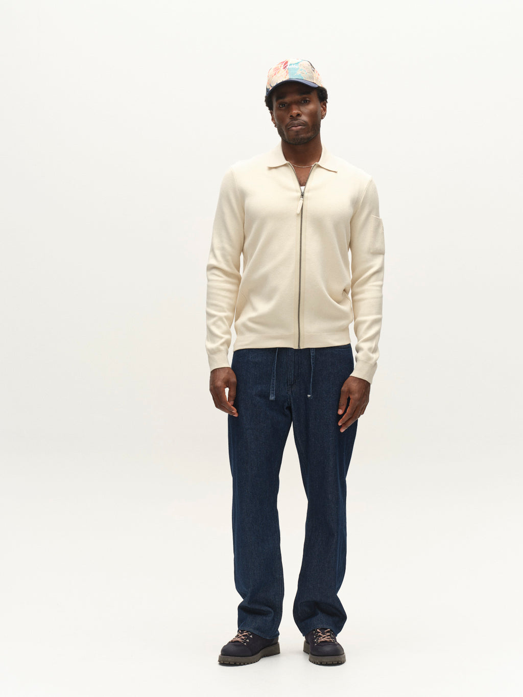 A man stands against a plain white background wearing the Gabba GABadriel Zip Lume LS Knit sweater in Birch, a regular-fit style crafted from a soft viscose blend knit with a full zip closure, paired with blue drawstring pants, black shoes, and a multicolored cap.