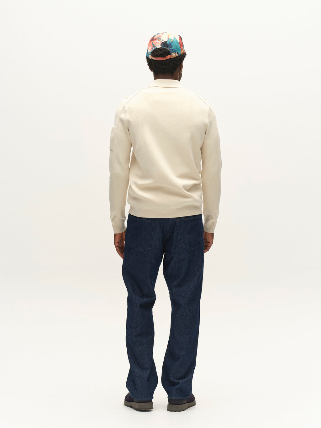 A person stands facing away, wearing the Gabba GABadriel Zip Lume LS Knit sweater in Birch—a cream-colored viscose blend knit—paired with blue jeans, black shoes, and a colorful patterned cap, against a plain white background.