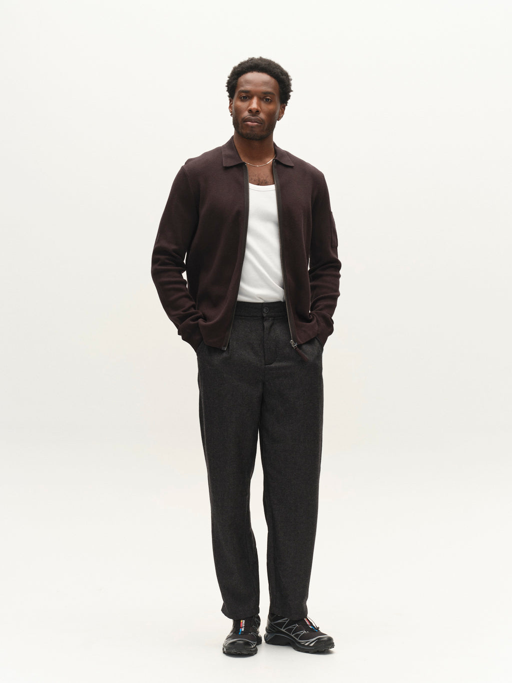 A man stands with his hands in his pockets, wearing the Gabba GABadriel Zip Lume LS Knit jacket in Meteorite over a white shirt, paired with dark gray pants and black sneakers, against a plain light background.
