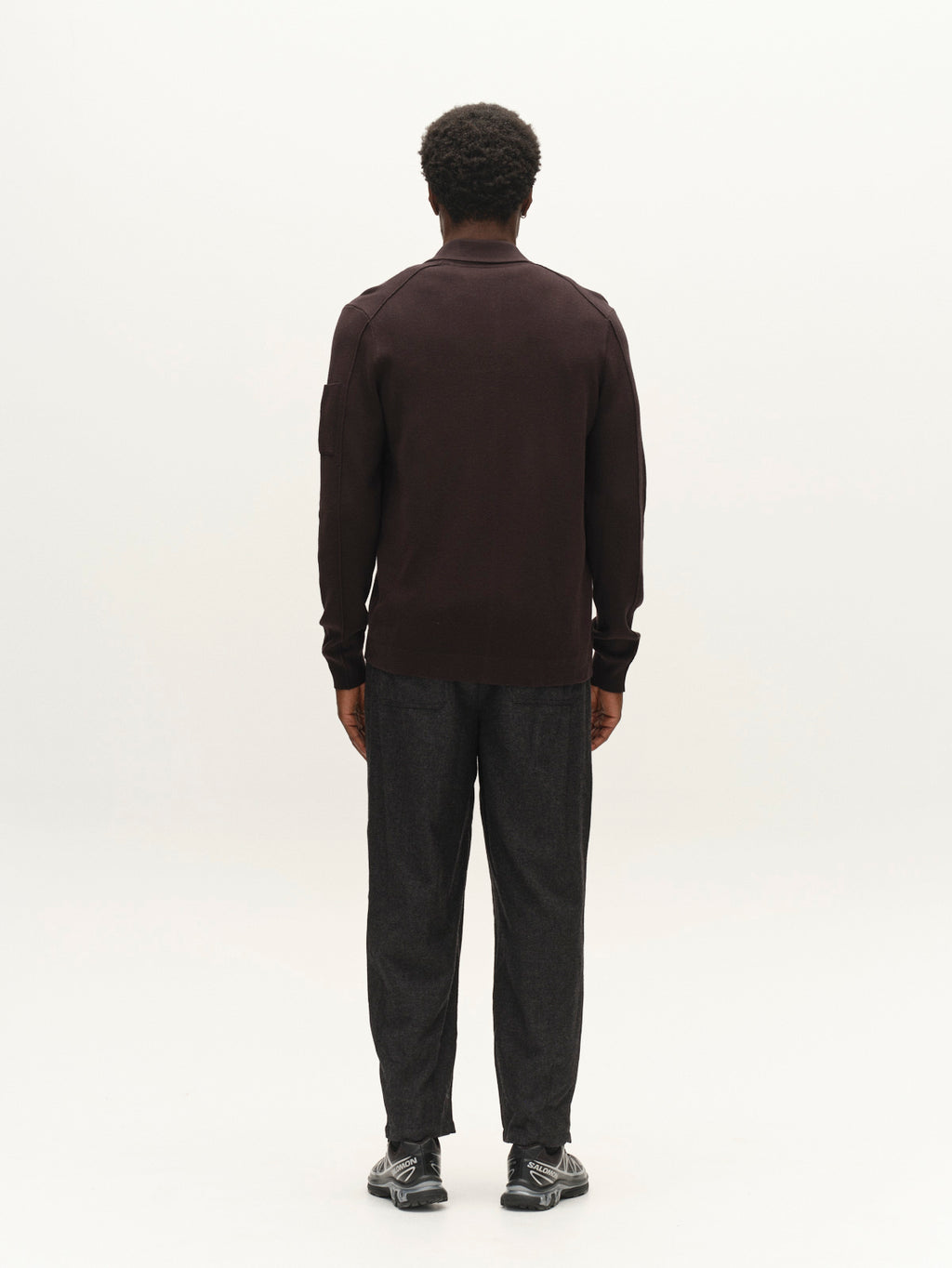 A person stands facing away from the camera, wearing the Gabba GABadriel Zip Lume LS Knit in Meteorite, paired with dark pants and black sneakers, against a plain white background.