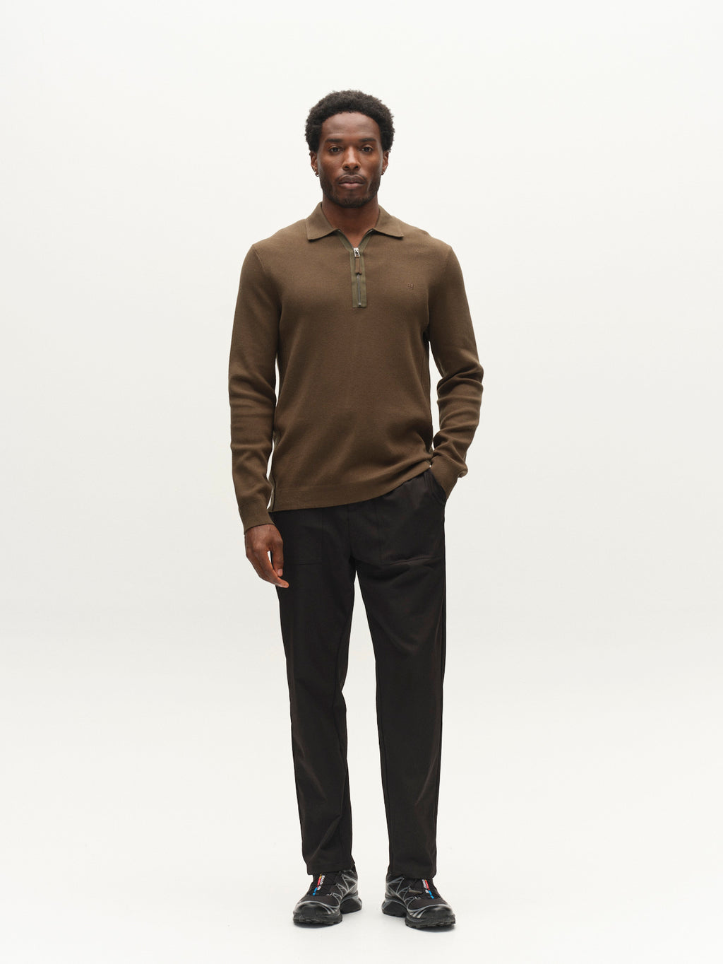 A man stands against a plain white background, wearing the Gabba GABadriel Half Zip Lume LS Knit in Forest Night, paired with black pants and black sneakers. He looks forward with a neutral expression, one hand in his pocket.