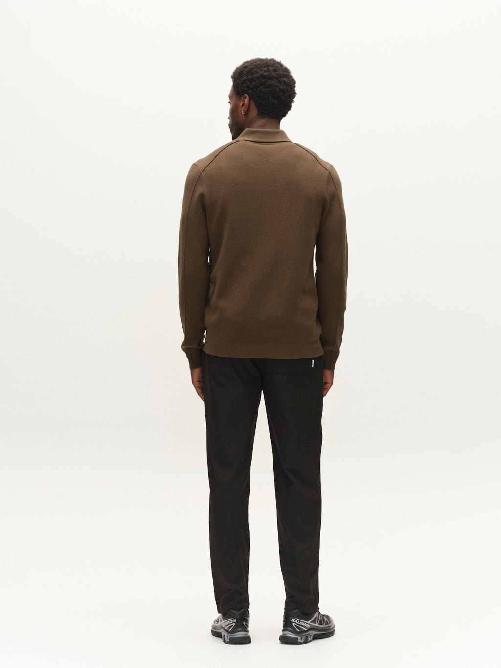 A man stands facing away, wearing the Gabba GABadriel Half Zip Lume LS Knit in Forest Night—a viscose blend half-zip sweater—paired with black pants and black shoes. The background is plain and light-colored.