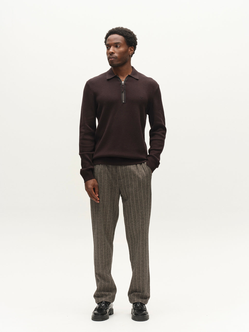 A man stands against a plain white background, wearing the Gabba GABadriel Half Zip Lume LS Knit in Meteorite, paired with gray pinstriped pants and black shoes. He looks slightly to his left with his hands relaxed by his sides.