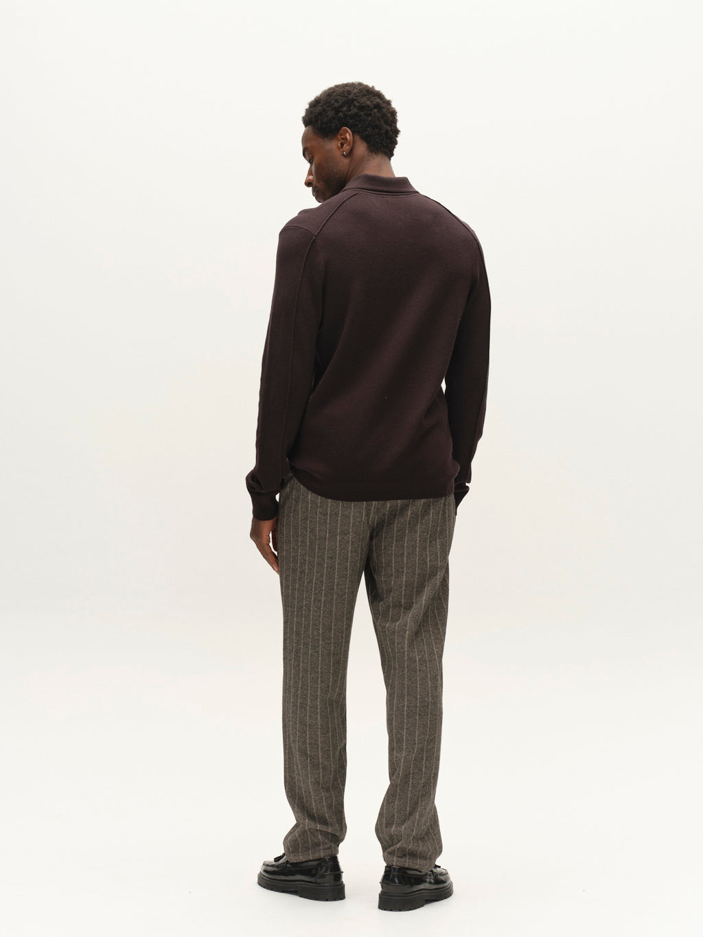 A man stands facing away, wearing the Gabba GABadriel Half Zip Lume LS Knit in Meteorite and gray pinstriped pants with black shoes, set against a plain light background.