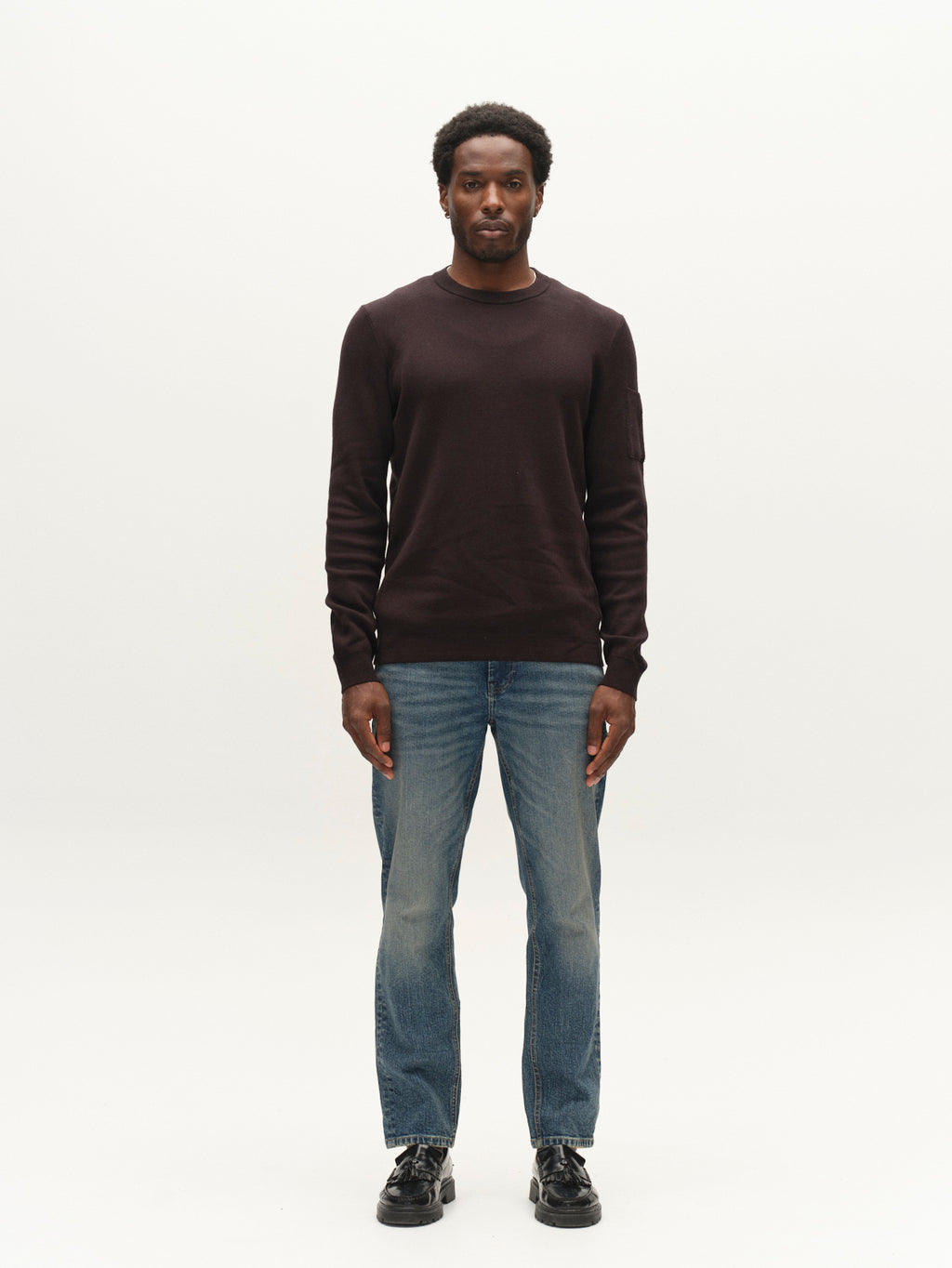 A man stands facing forward, wearing the Gabba GABadriel O-Neck Lume LS Knit sweater in Meteorite, paired with blue jeans and black shoes, against a plain white background.