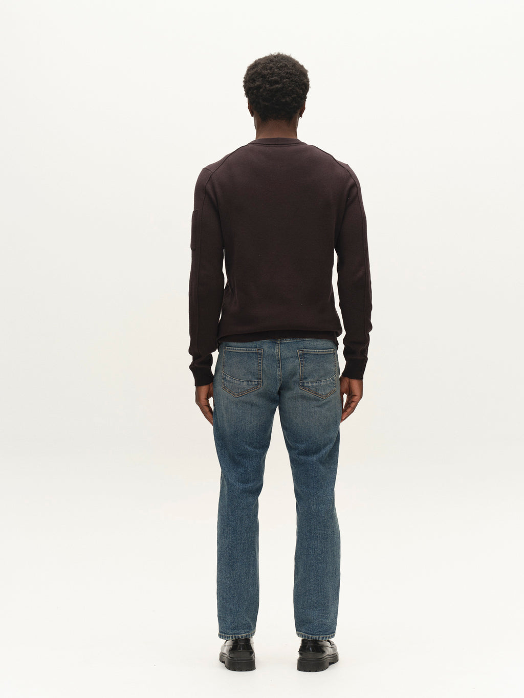 Wearing the Gabba GABadriel O-Neck Lume LS Knit in Meteorite, a person with short curly hair stands facing away against a plain white background. The look is completed with blue jeans and black shoes.
