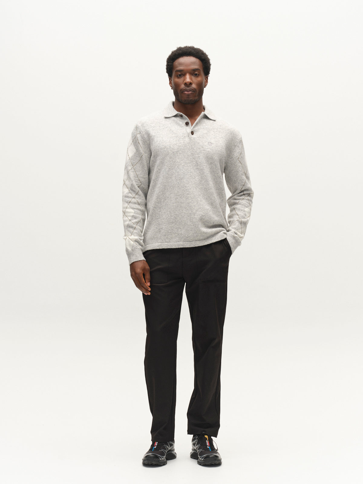 A man stands facing forward wearing the Gabba GABpietro Polo Argyle LS Knit in Light Grey Mel, paired with black pants and black sneakers, against a plain white background.