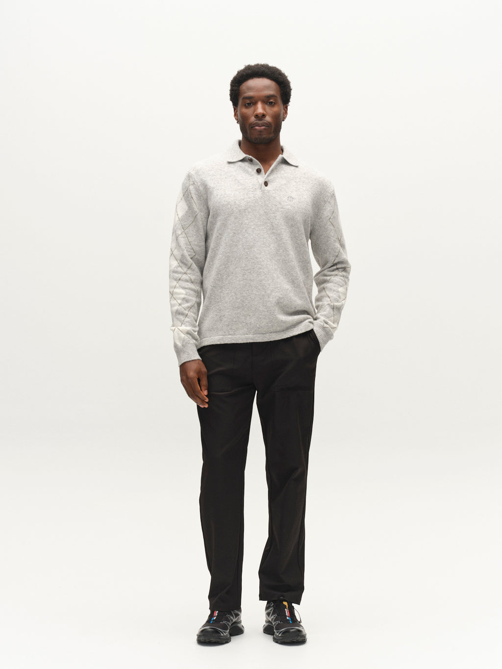 A man stands facing forward wearing the Gabba GABpietro Polo Argyle LS Knit in Light Grey Mel, paired with black pants and black sneakers, against a plain white background.