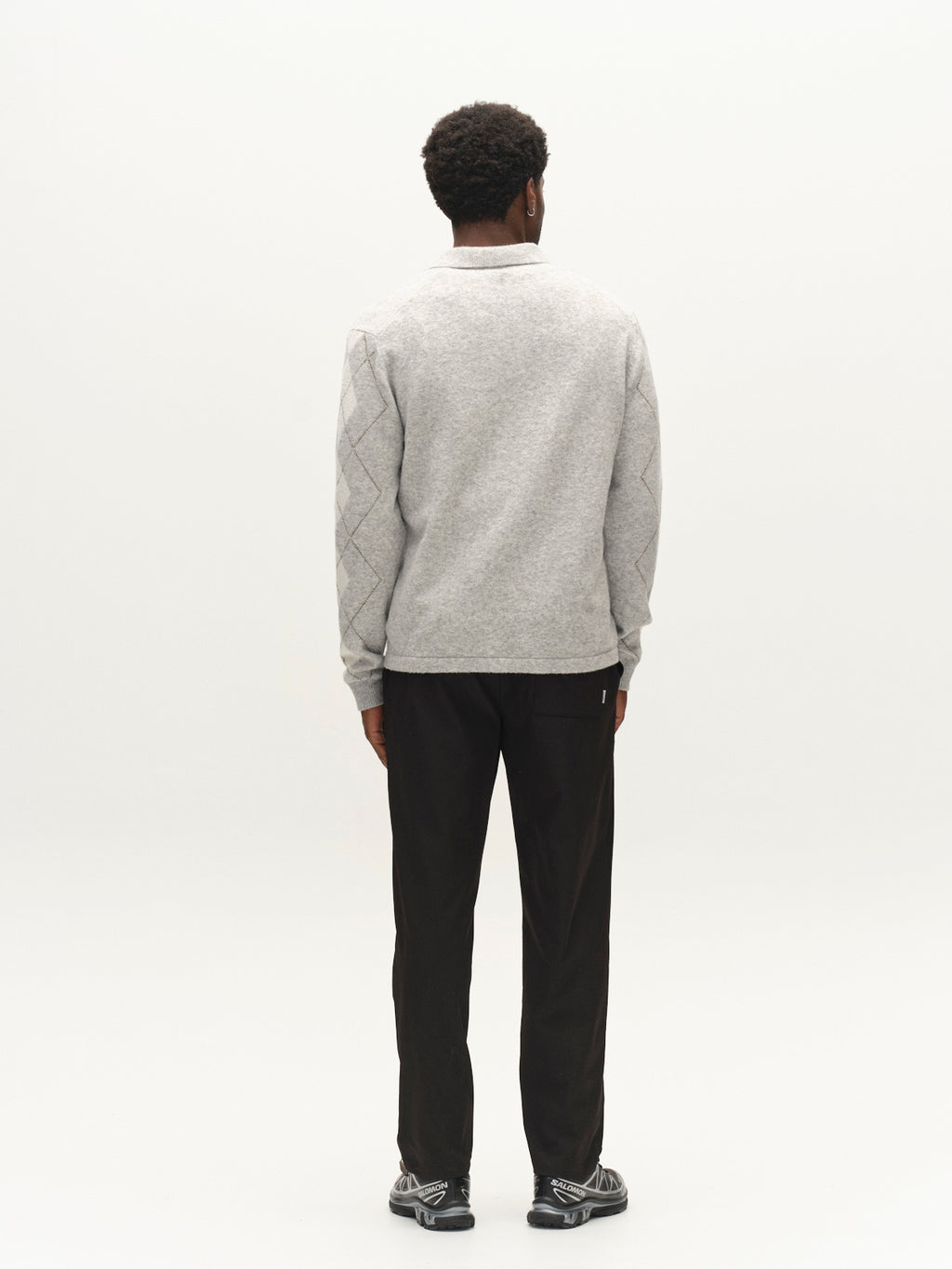 A man stands facing away from the camera, wearing the Gabba GABpietro Polo Argyle LS Knit in Lt. Grey Mel—a modern sweater with a diamond pattern on the sleeves—paired with black pants and black-and-gray sneakers, set against a plain white background.