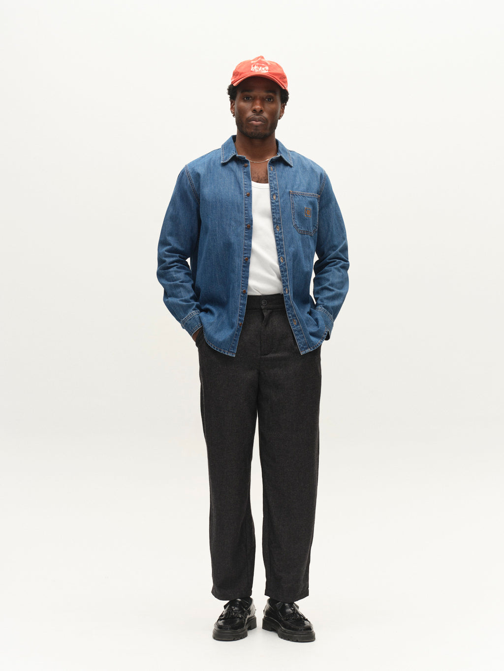 A man stands facing forward, wearing a red cap, the Gabba GABsven Reg Wish LS Shirt in mid blue denim over a white T-shirt, dark pants, and black shoes against a plain white background. This versatile wardrobe essential from Gabba adds timeless style to his look.